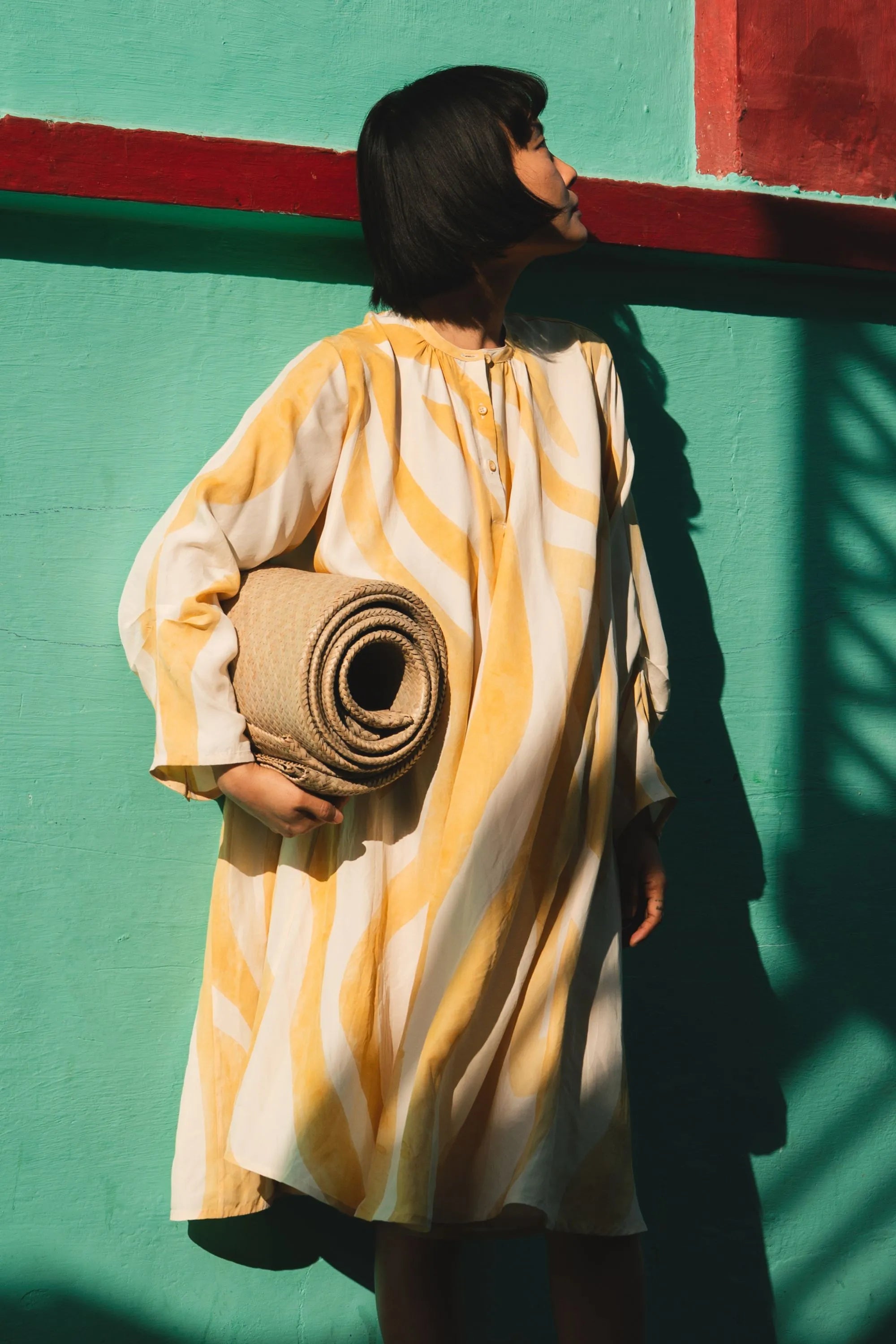 A woman wearing mango yellow dress and holding a rolled-up mat against a green wall with red accents