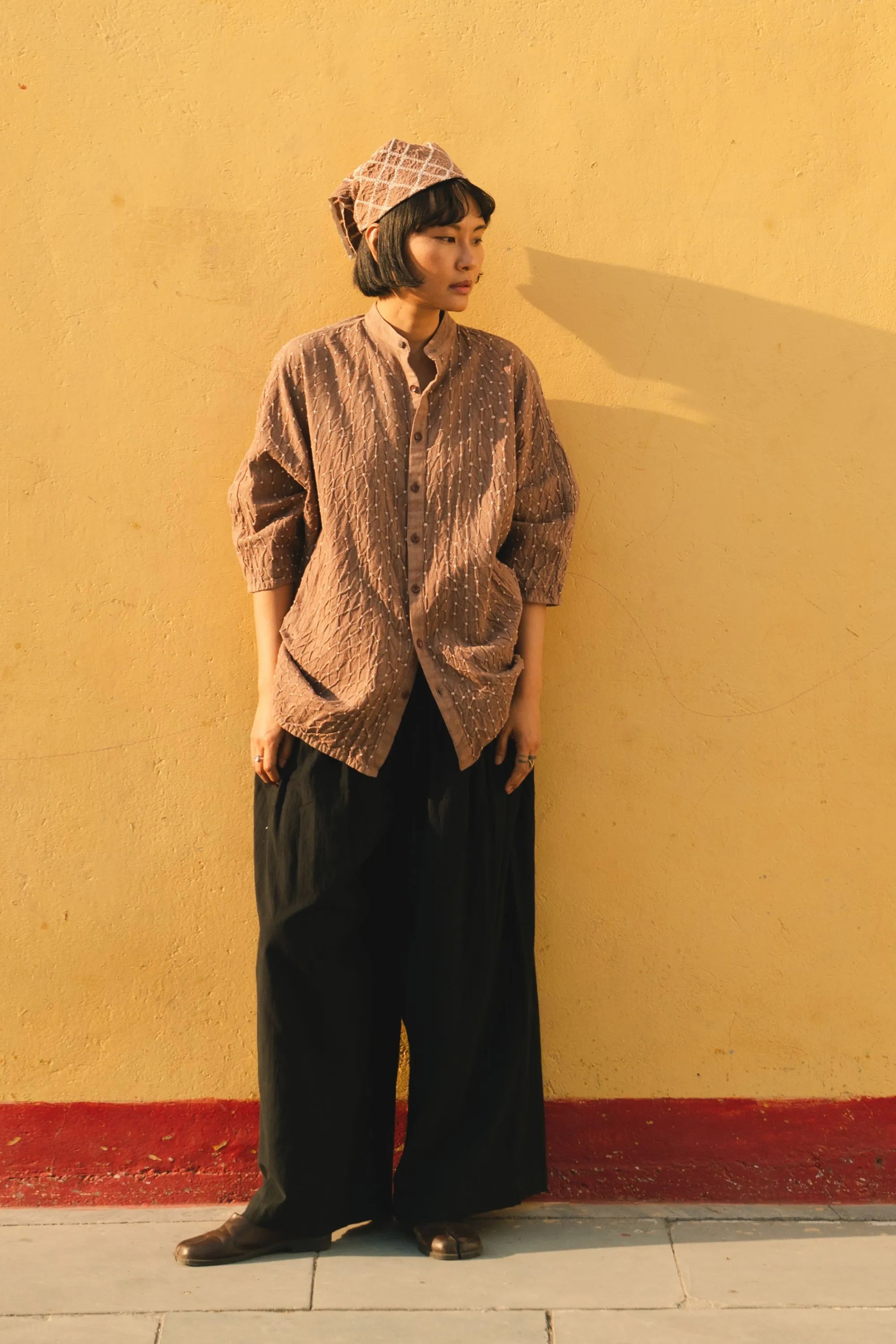 A woman wearing a old rose shirt and black pants against a yellow wall.