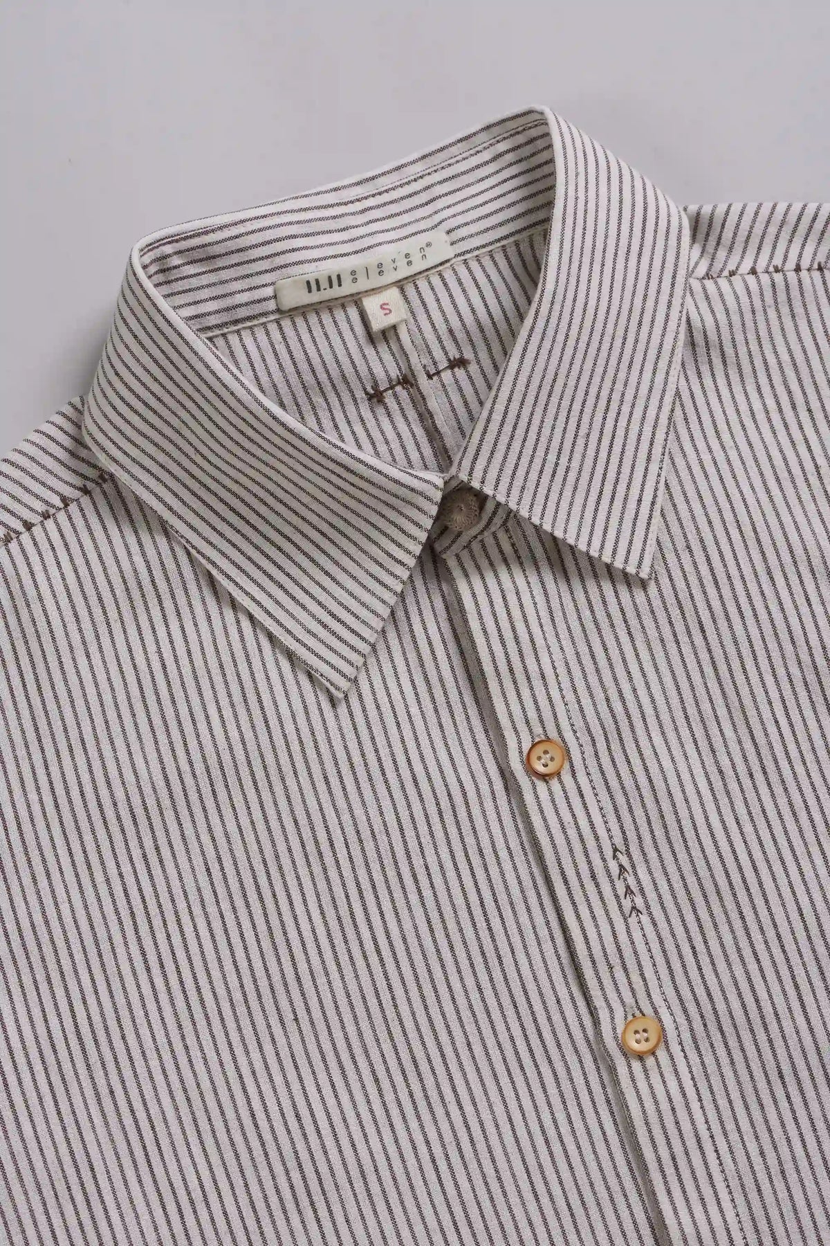 Close-up of a collared button-up shirt with thin vertical dark pinstripes on a light beige or off-white base, featuring light brown buttons.