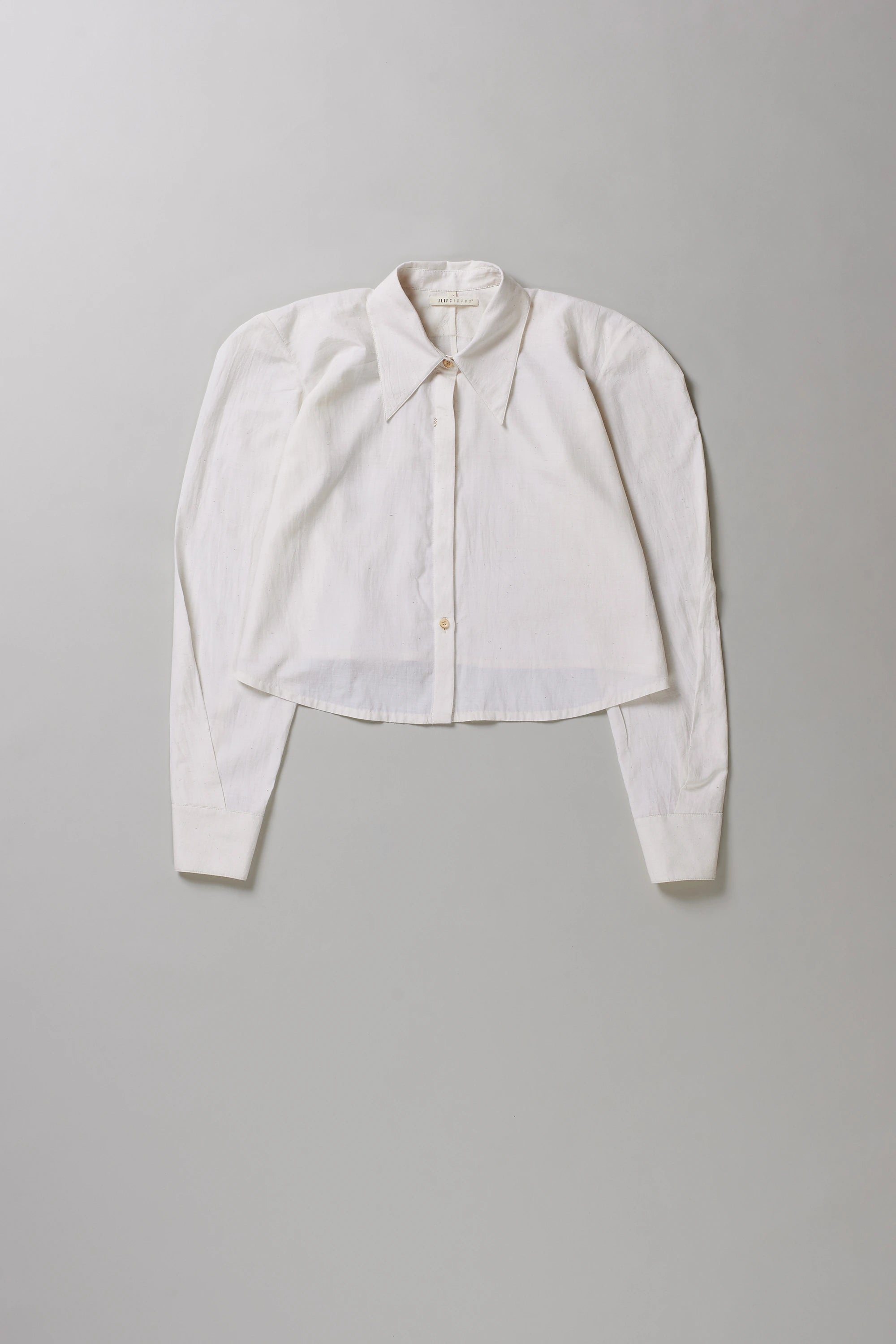 A cropped long-sleeve button-up shirt in off-white, laid flat against a neutral light gray background.