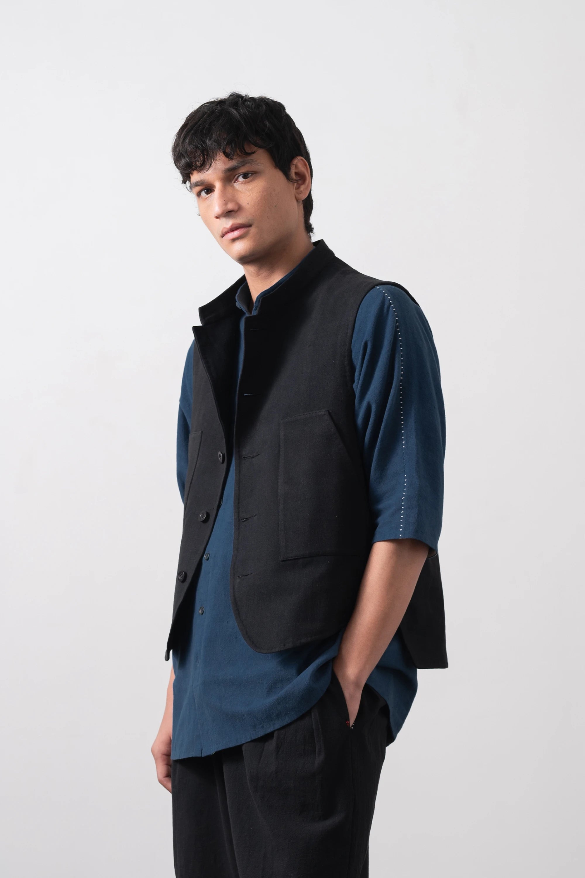 A man wearing a black sleeveless vest over a dark blue short-sleeve shirt and black trousers, standing against a light grey studio background.