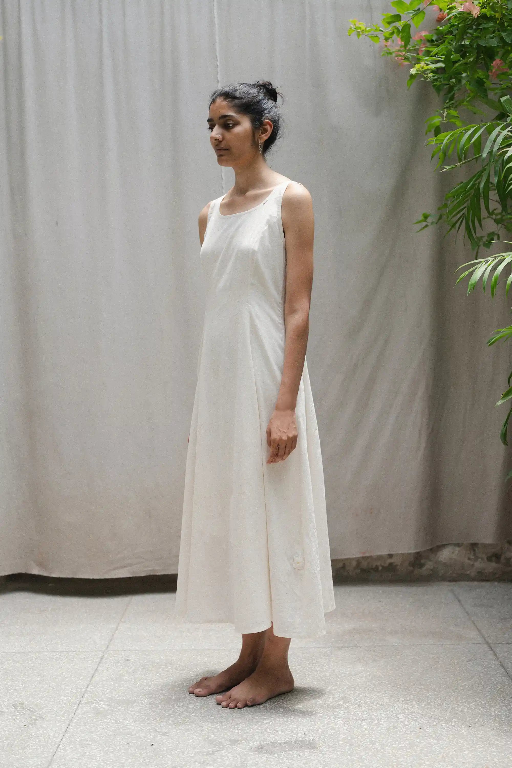 Woman wearing a ecru dress standing against a plain background with plants.