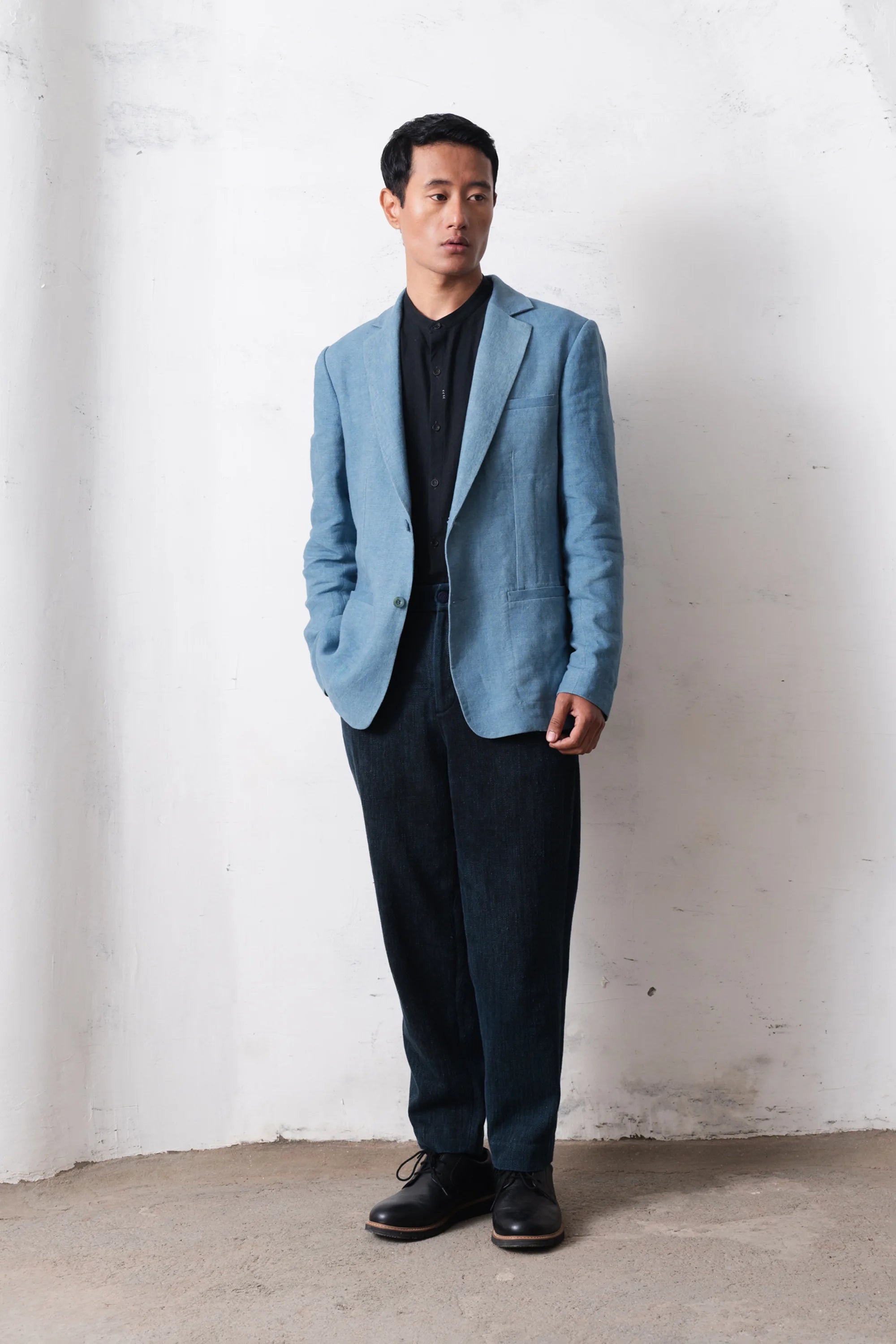Man wearing a light blue blazer over a black t-shirt and pants against a light grey wall