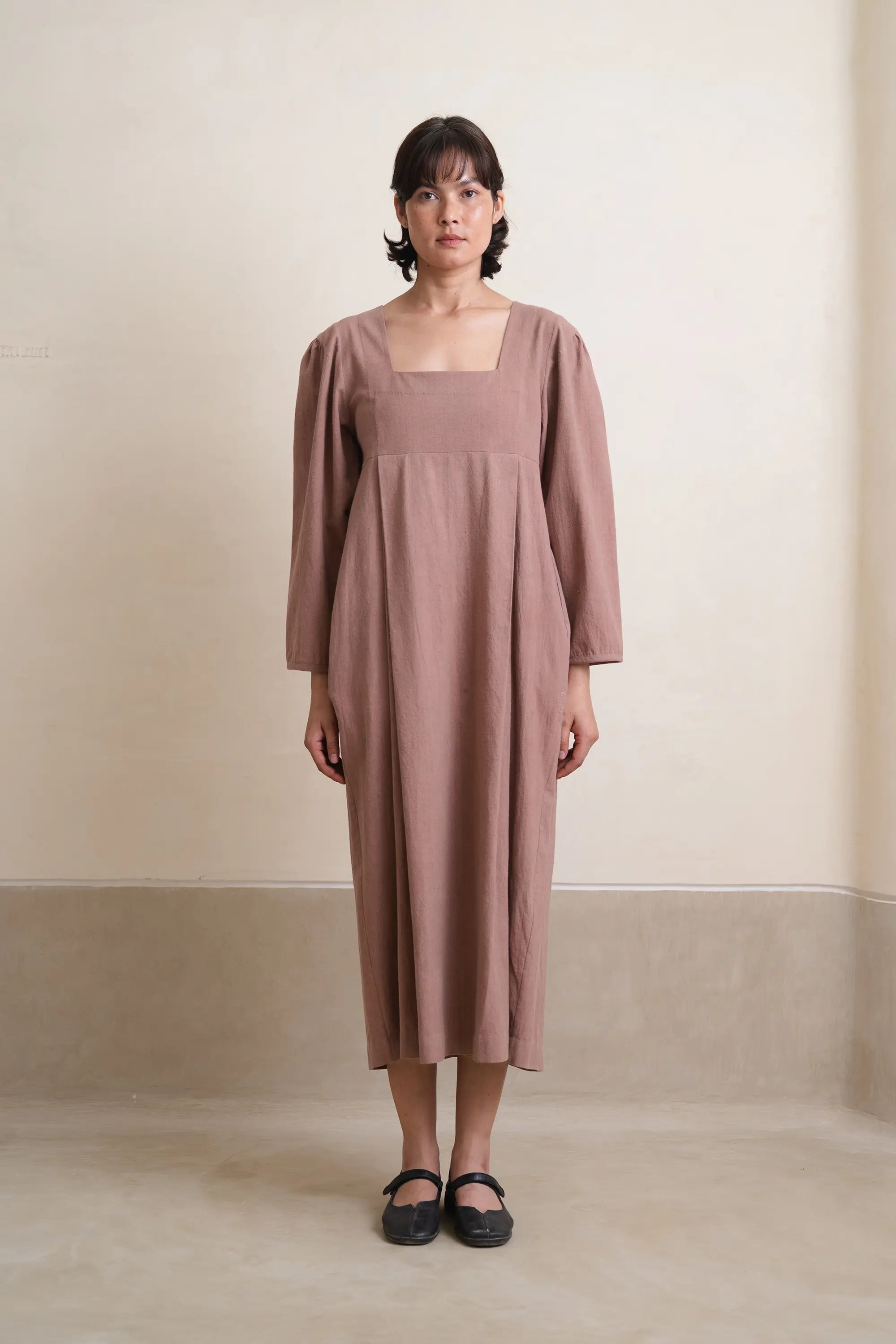 Woman wearing a long, light brown dress against a plain background