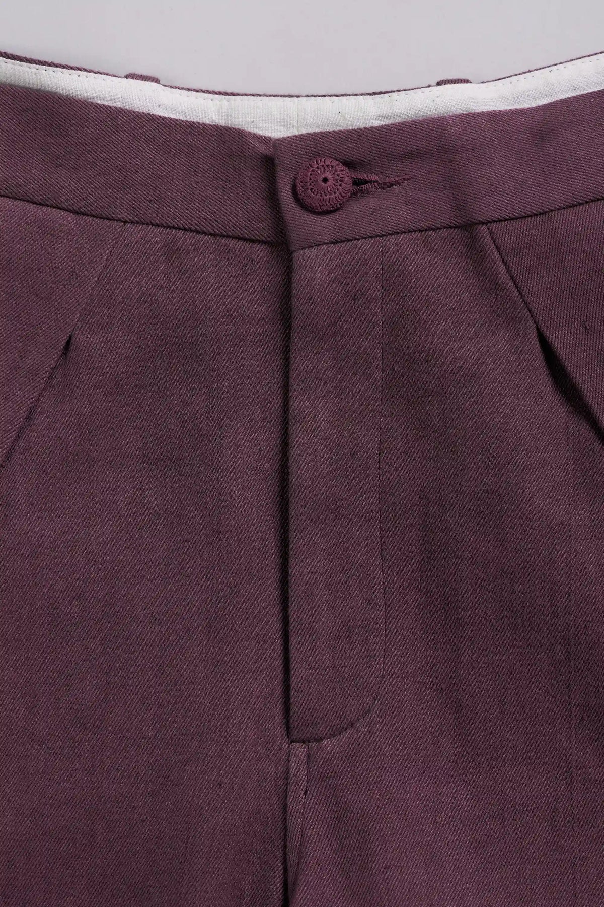 Close-up of a pair of trousers in a purple fabric, showing the button closure, waistband, and front pleats.