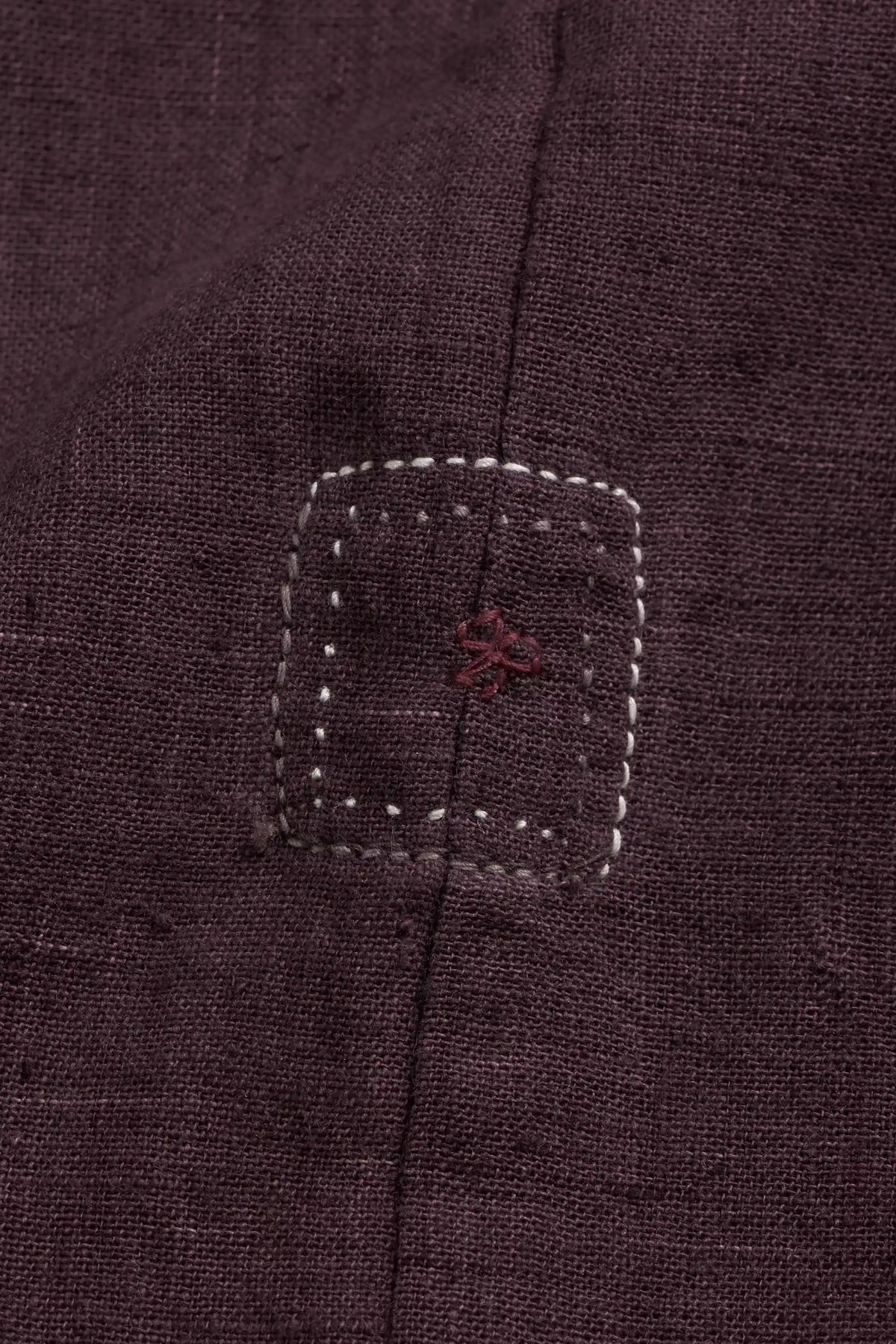 Close-up of a purple red textured fabric with stitched in the center, featuring a small red embroidered cross motif and white contrast stitching.
