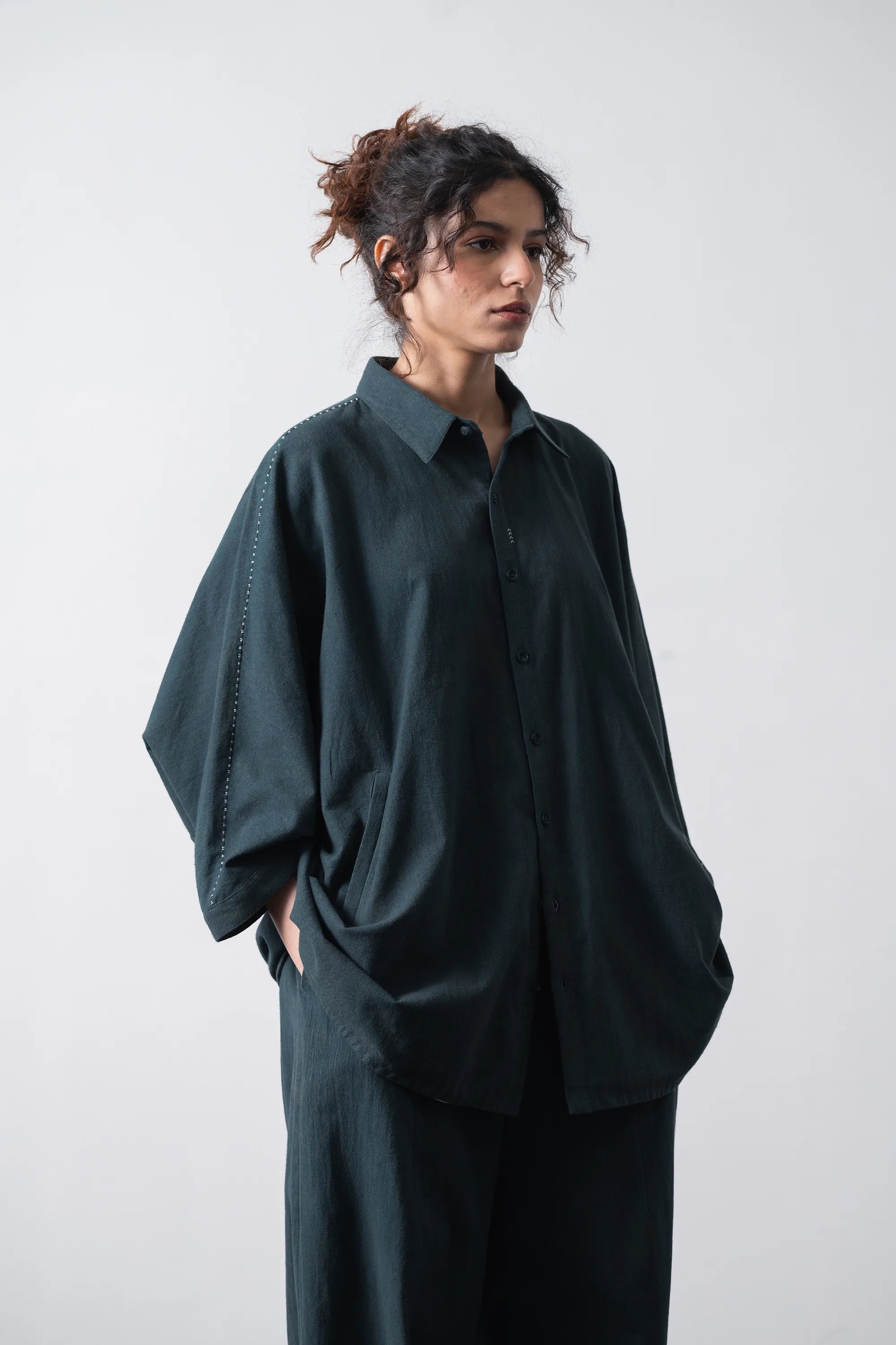 A woman wearing an oversized black indigo button-up shirt and matching wide-leg trousers, standing against a light grey studio background.