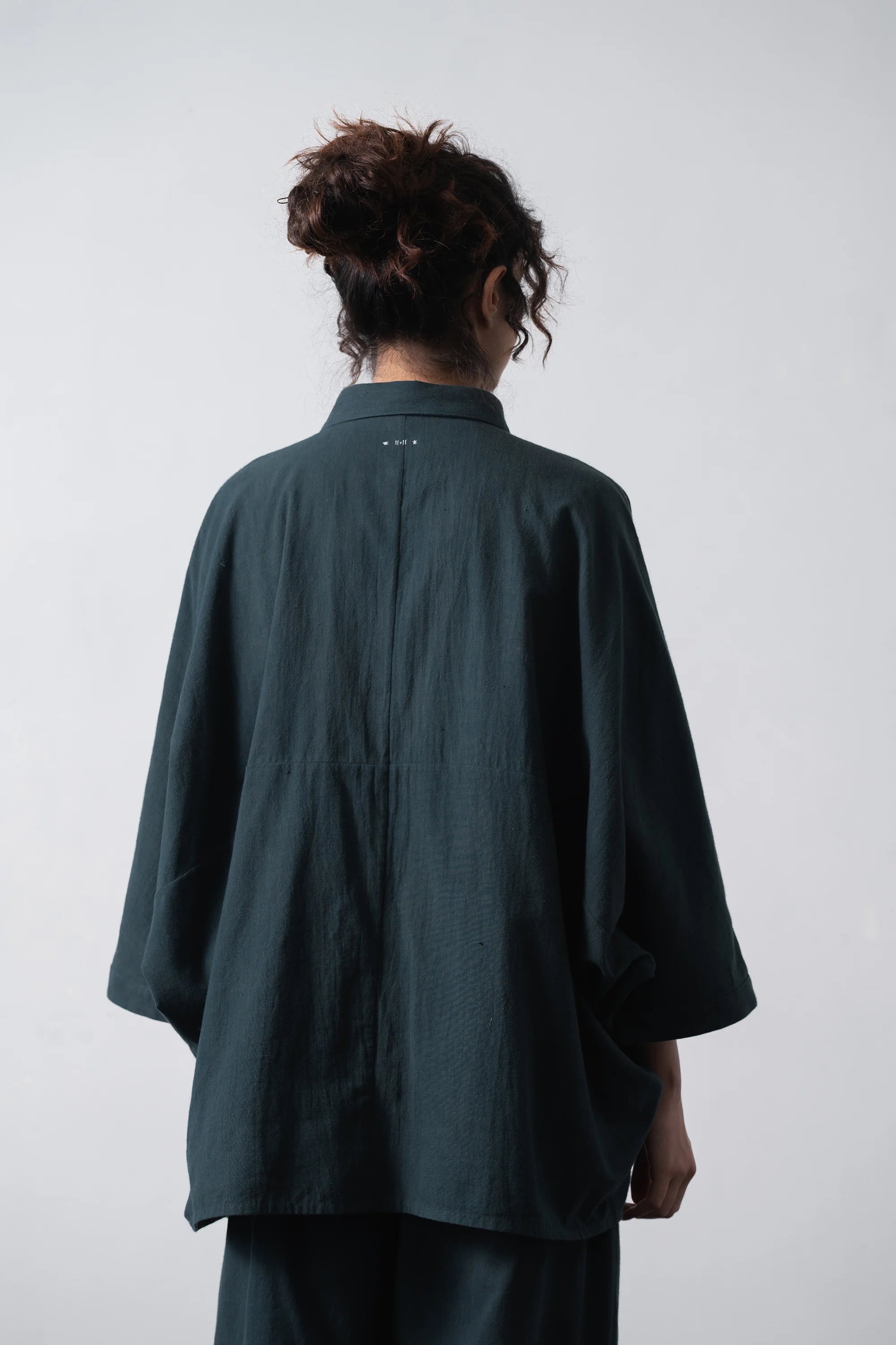 A woman model shown from the back wearing an oversized black indigo shirts, against a plain light grey studio background.