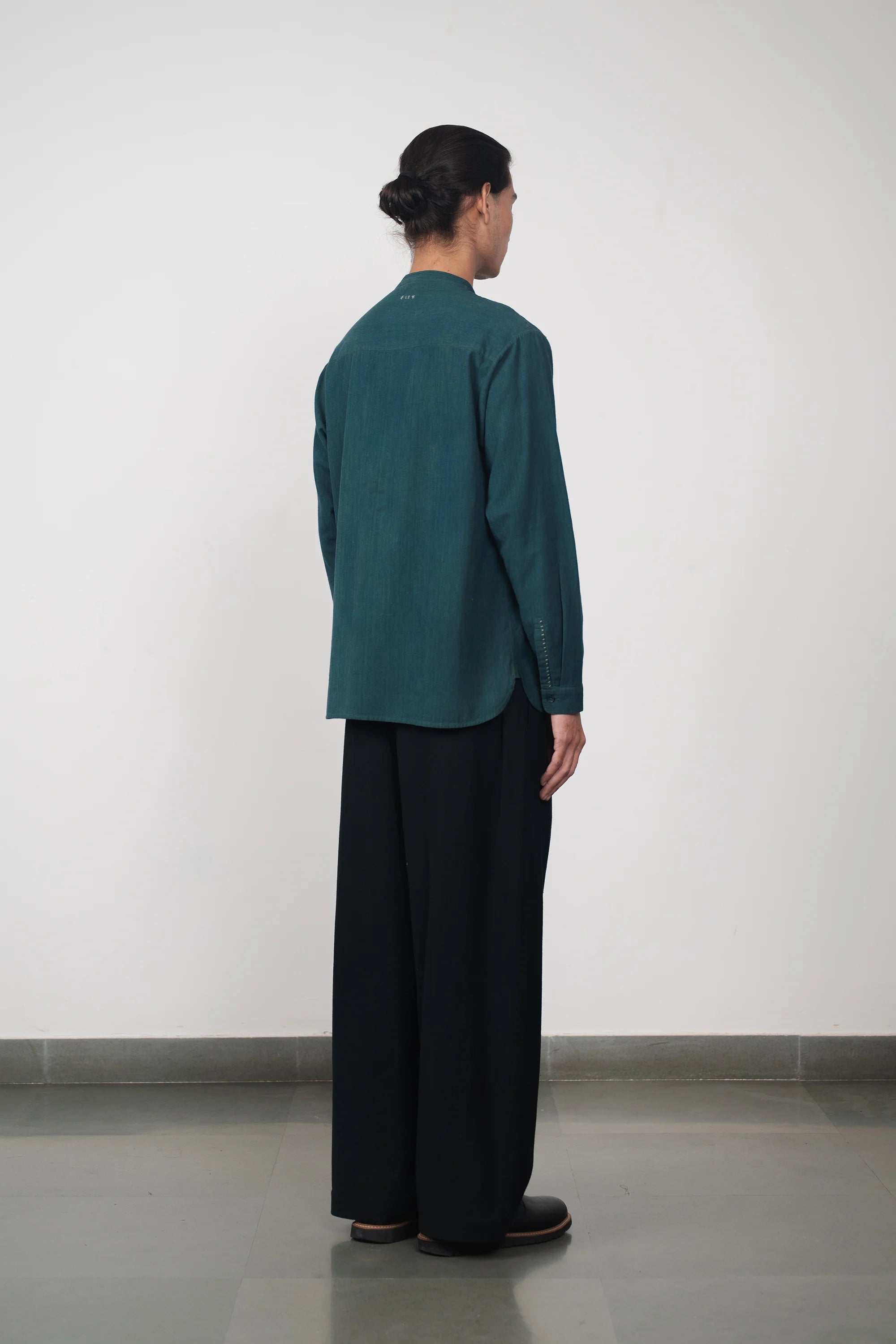 Side shot of a person wearing a green shirt and black pants standing against a light grey wall