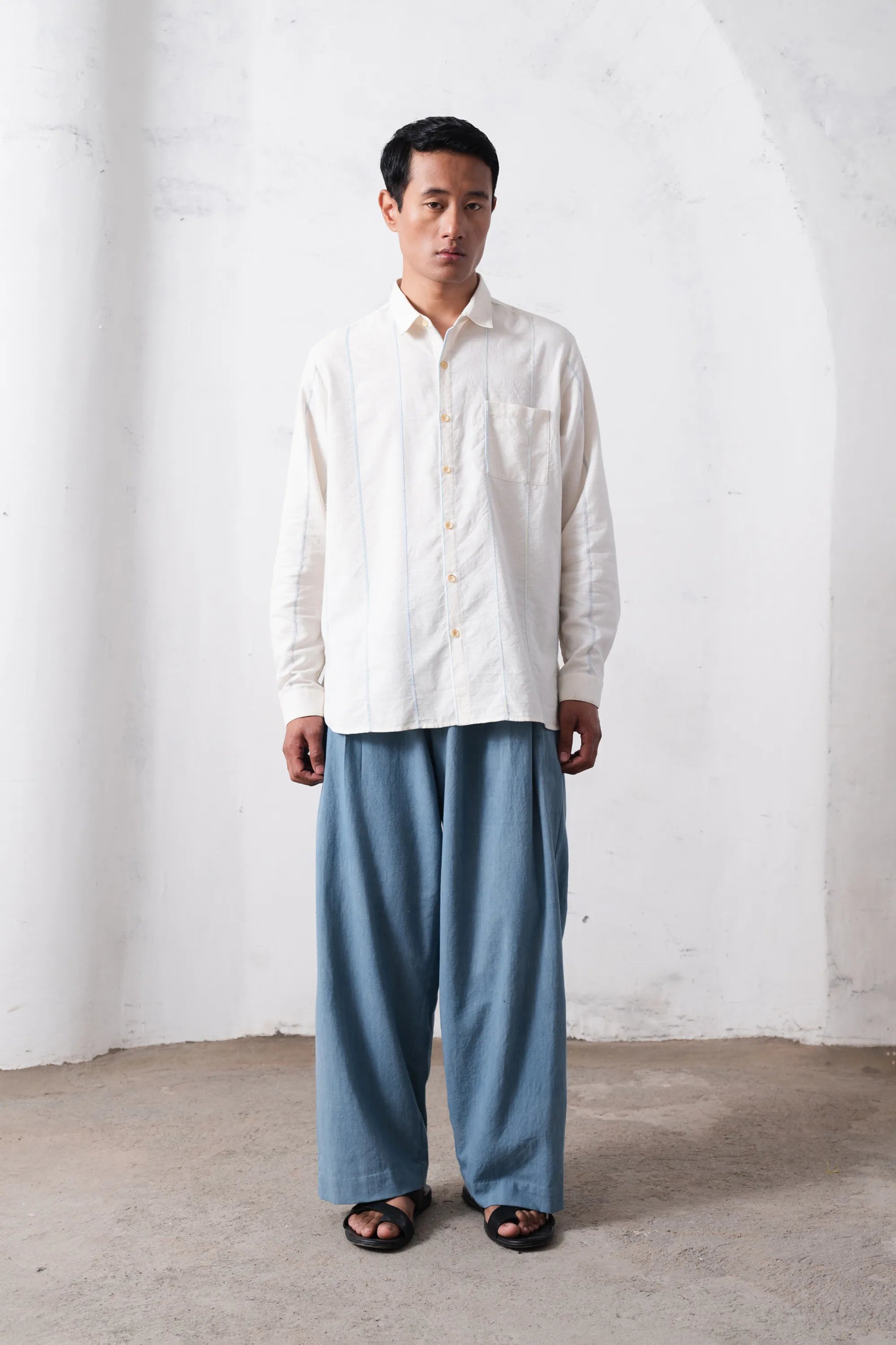 Man wearing a white shirt and blue pants standing against a plain light grey wall