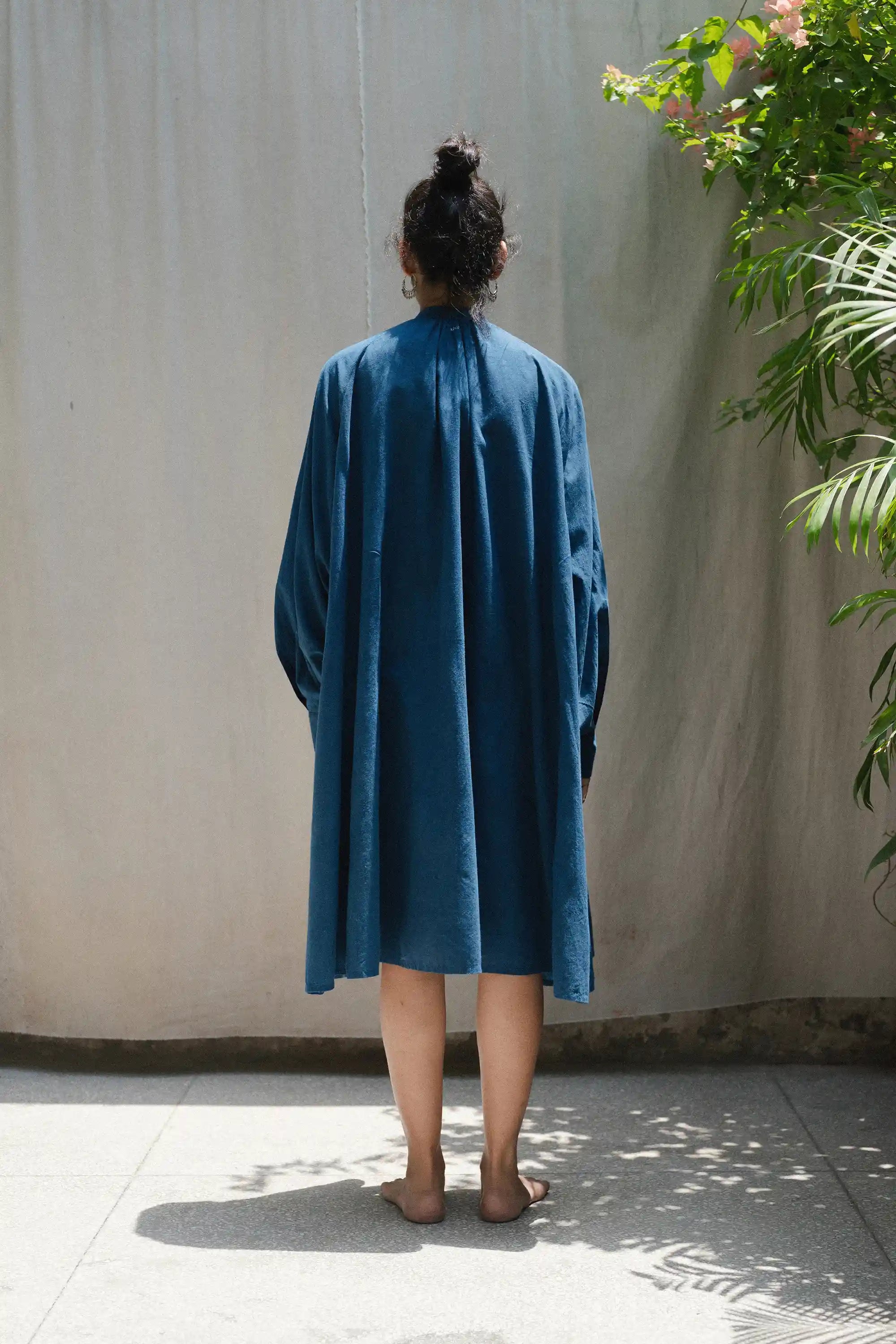 Back shot of a person wearing a blue dress standing against a plain wall with plants around.