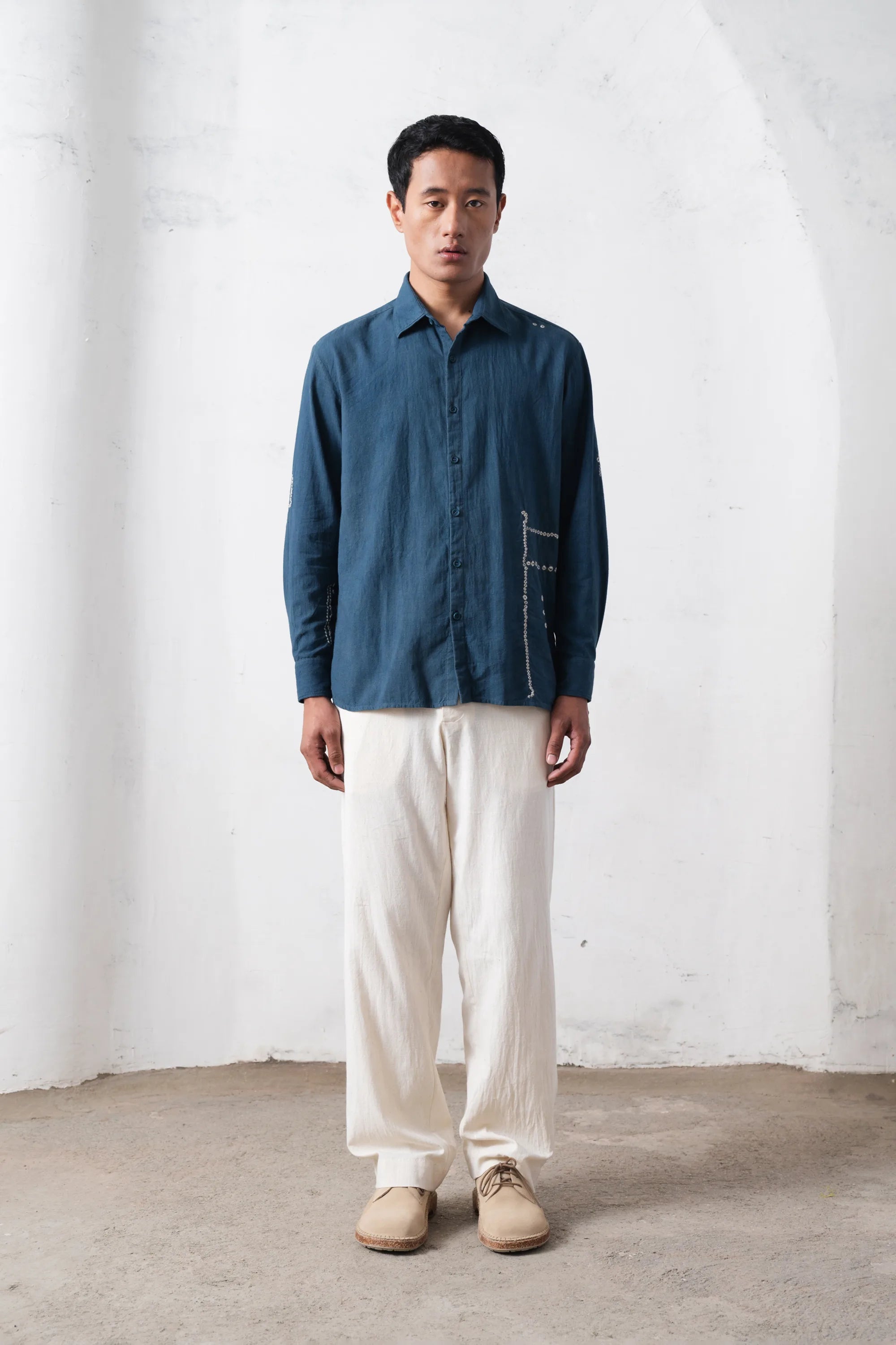 Man wearing a blue shirt and white pants standing against a white wall