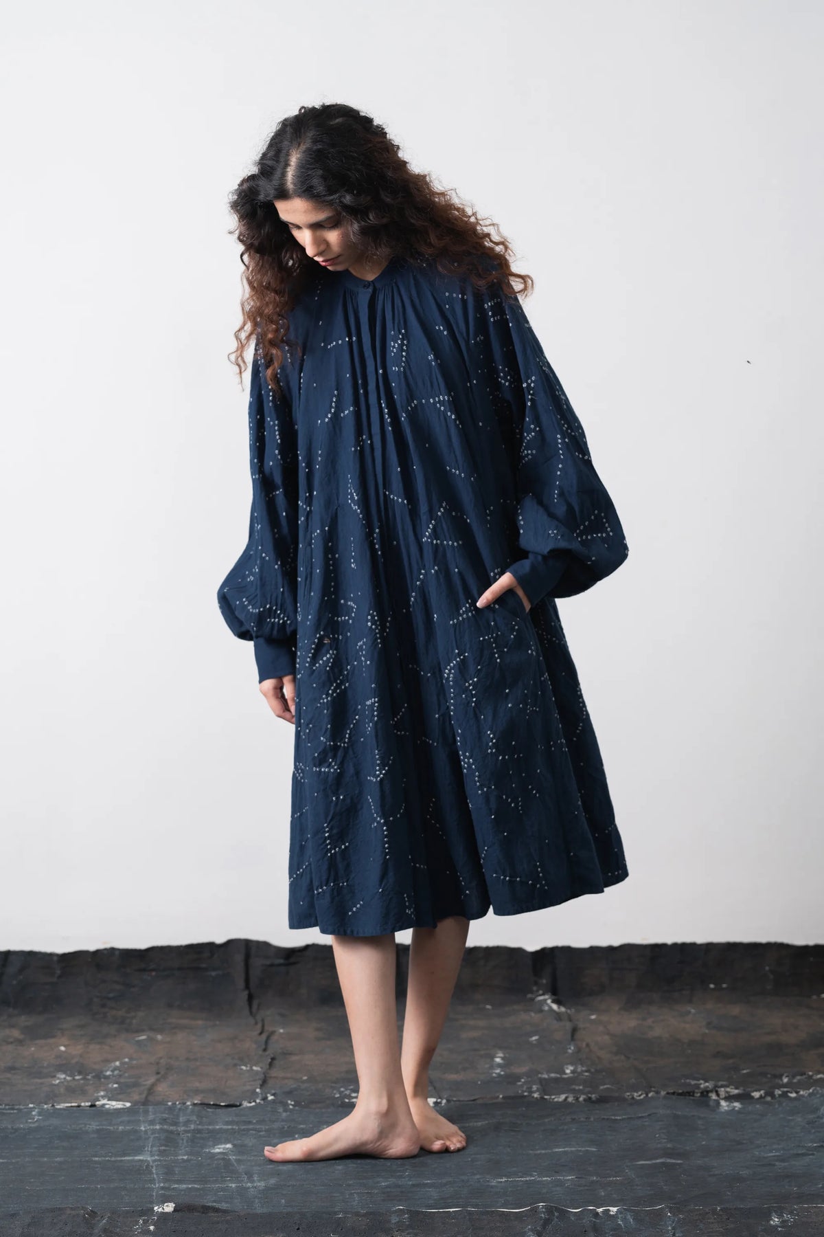 A woman stands barefoot against a plain off-white wall, wearing a loose space blue dress with a bandhani artwork and voluminous long sleeves.
