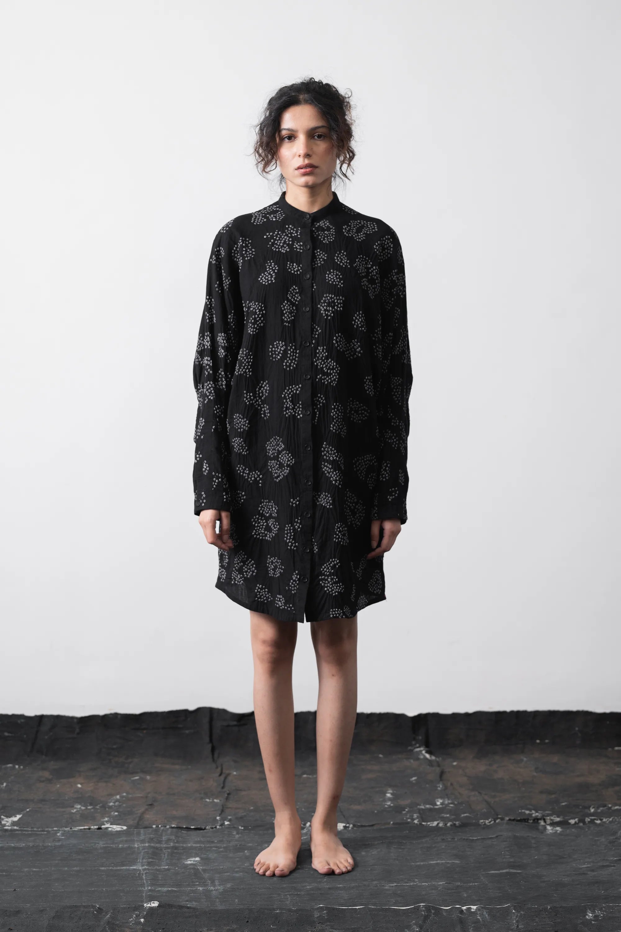 A model stands barefoot against a white wall and dark floor, wearing a black long-sleeve shirt dress with a grey floral pattern.