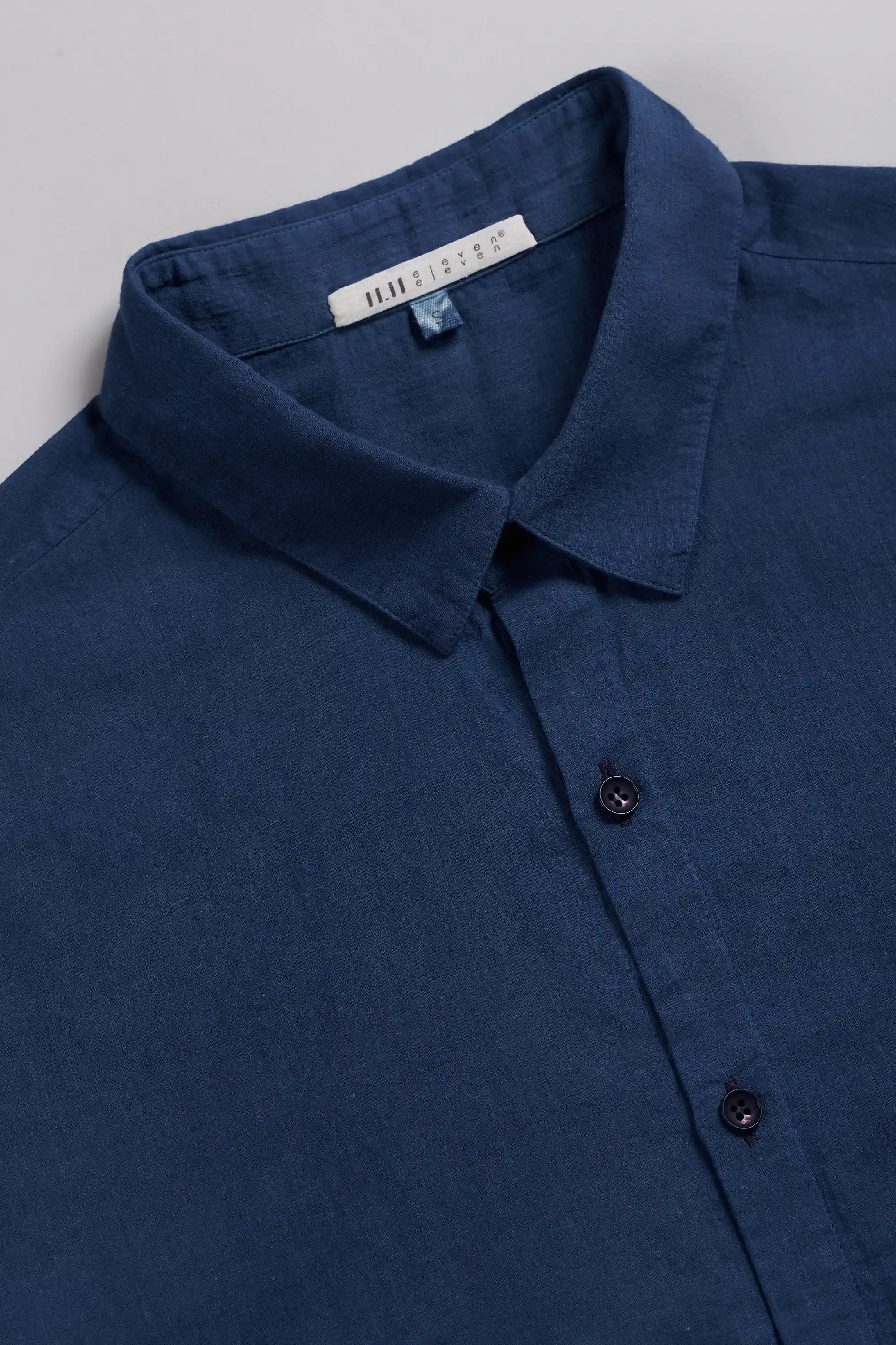 A close-up of a medium indigo shirt collar with a visible brand label, showing the textured fabric and dark buttons.