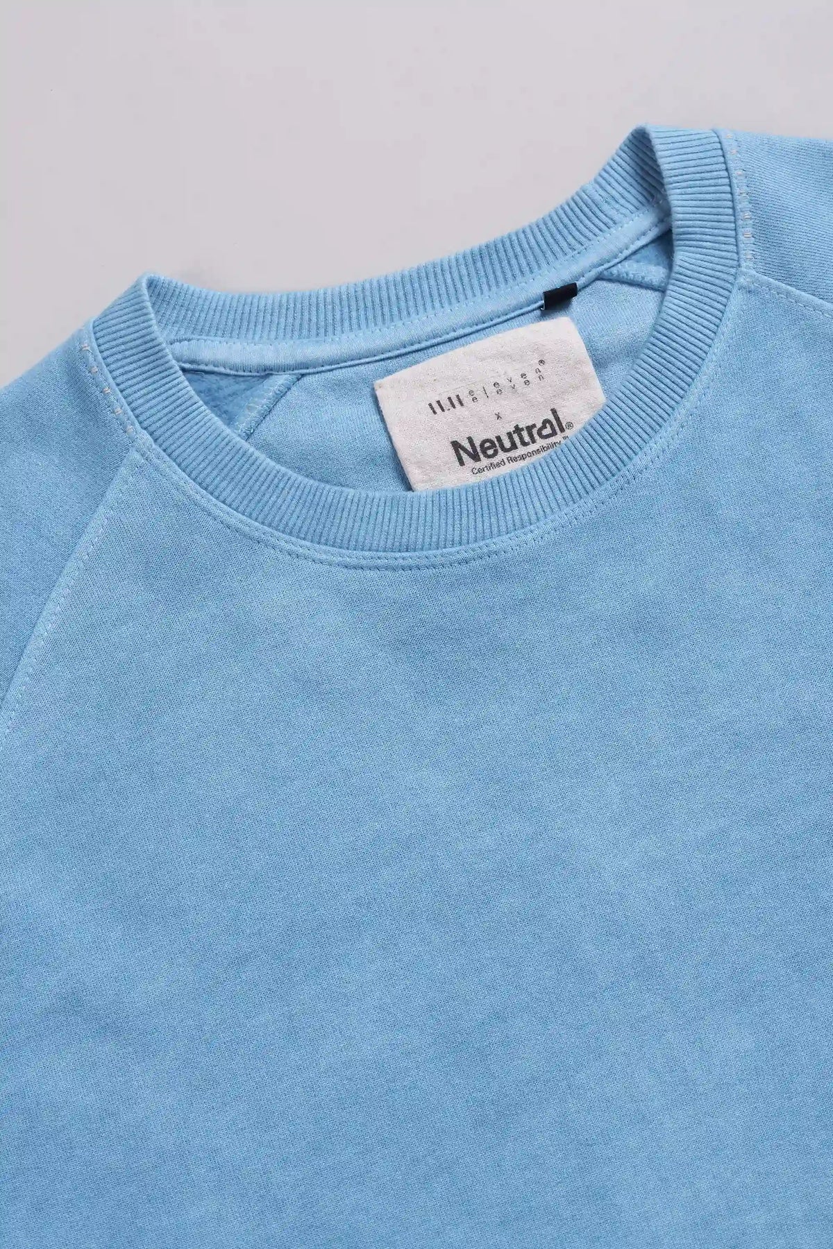A close-up of a light blue crewneck sweatshirt, showing the ribbed collar and a white brand label that reads 'Neutral'.