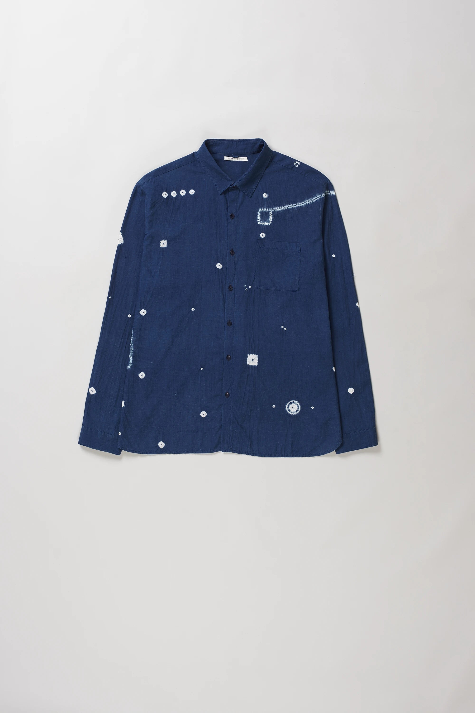 A medium indigo long-sleeve button-up shirt with white shibori details, laid flat against a light grey background.