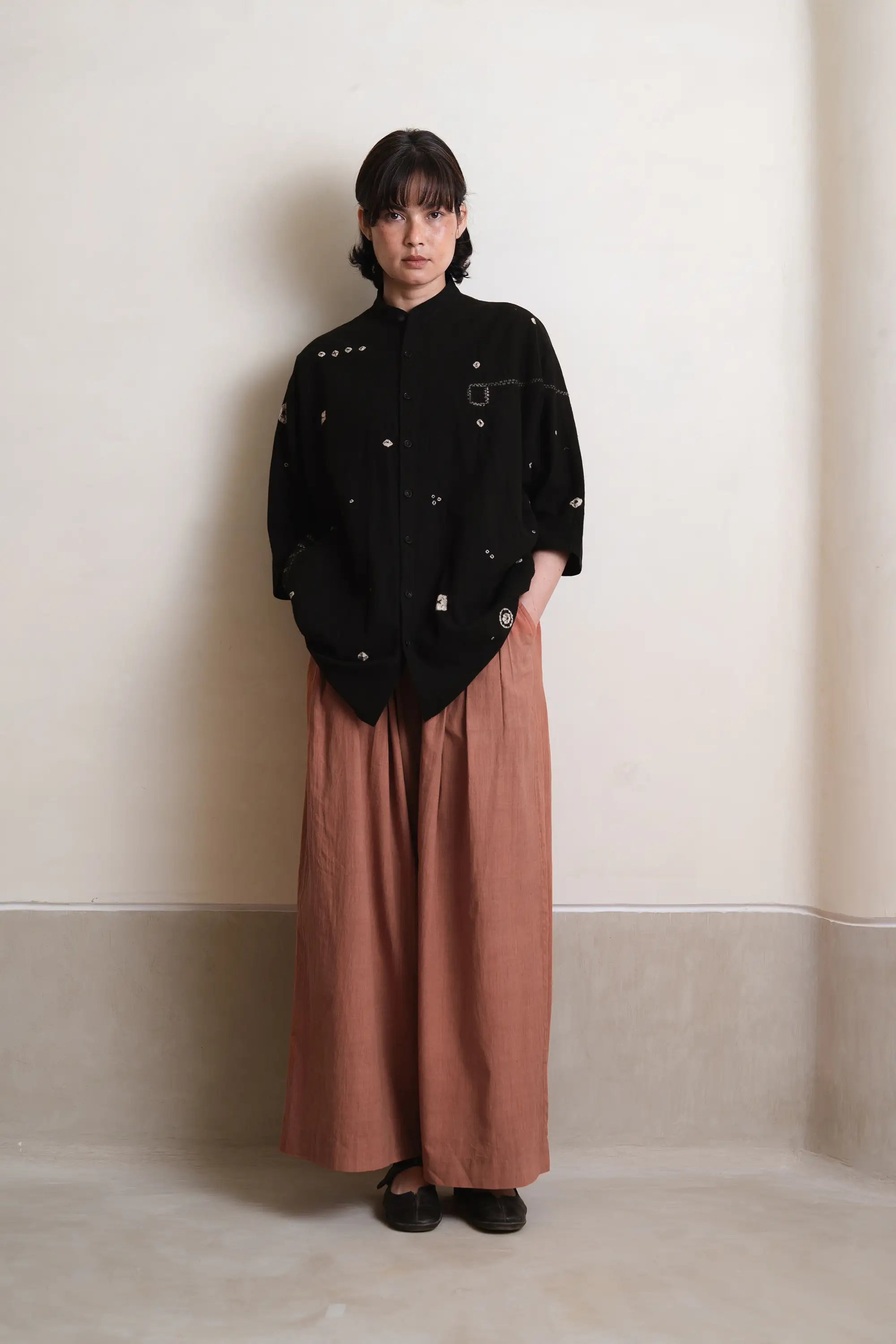 Person wearing a black jacket with white patterns and brown pants against a plain background