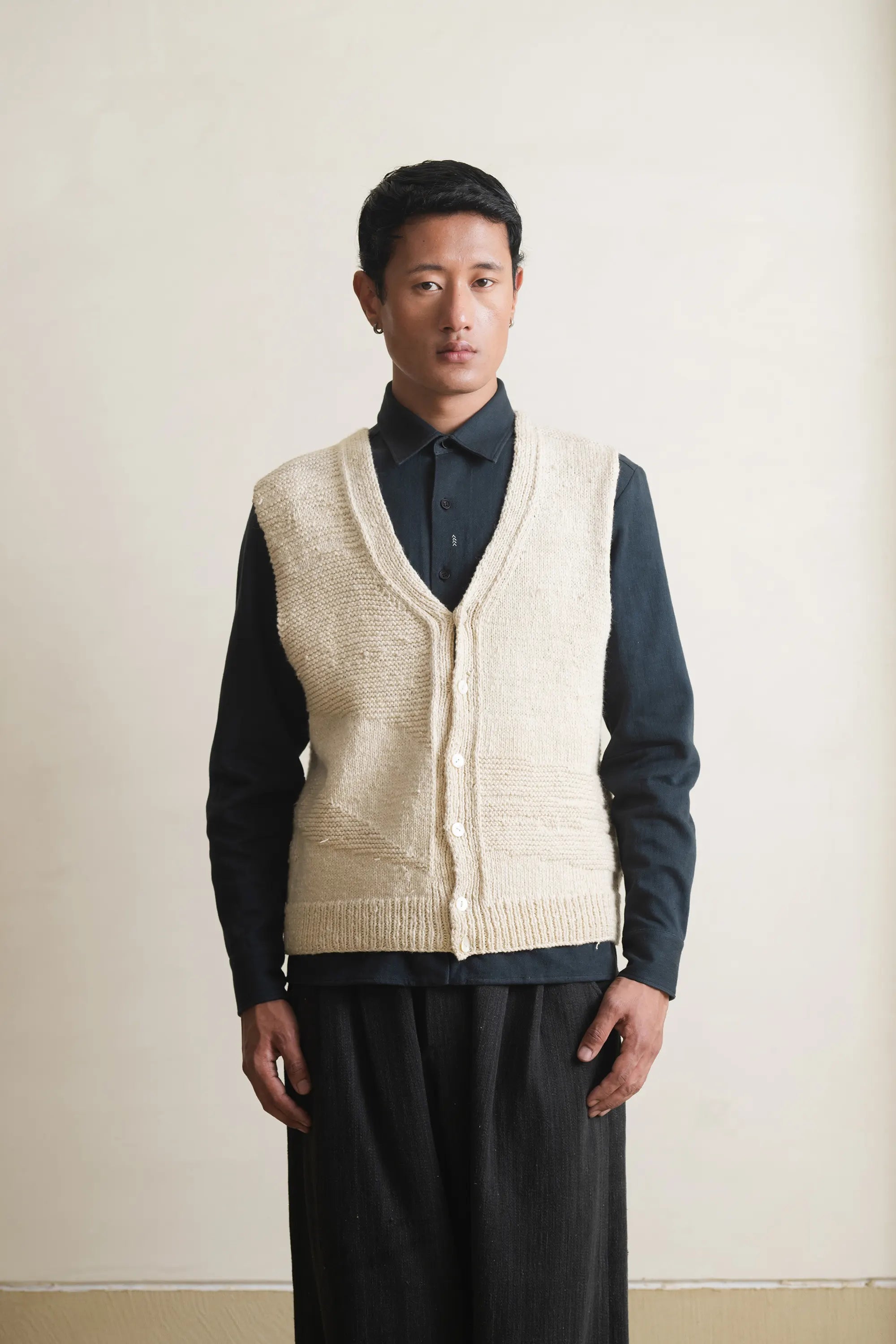 Man wearing a cream knitted vest over a black shirt against a light grey background