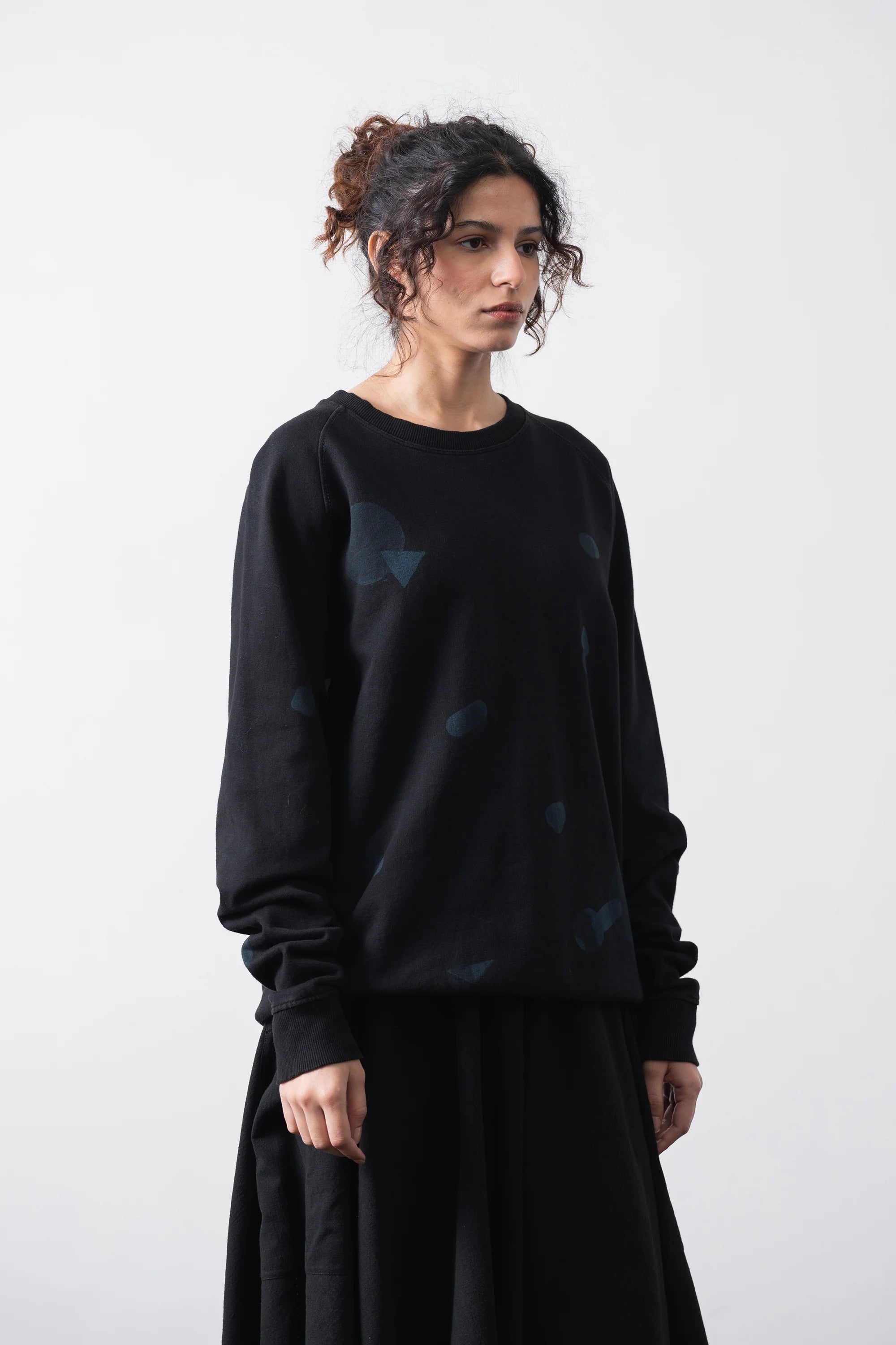 A woman model a black crewneck sweatshirt with subtle blue geometric patterns, paired with a black sweatshirt, against a plain light grey background.