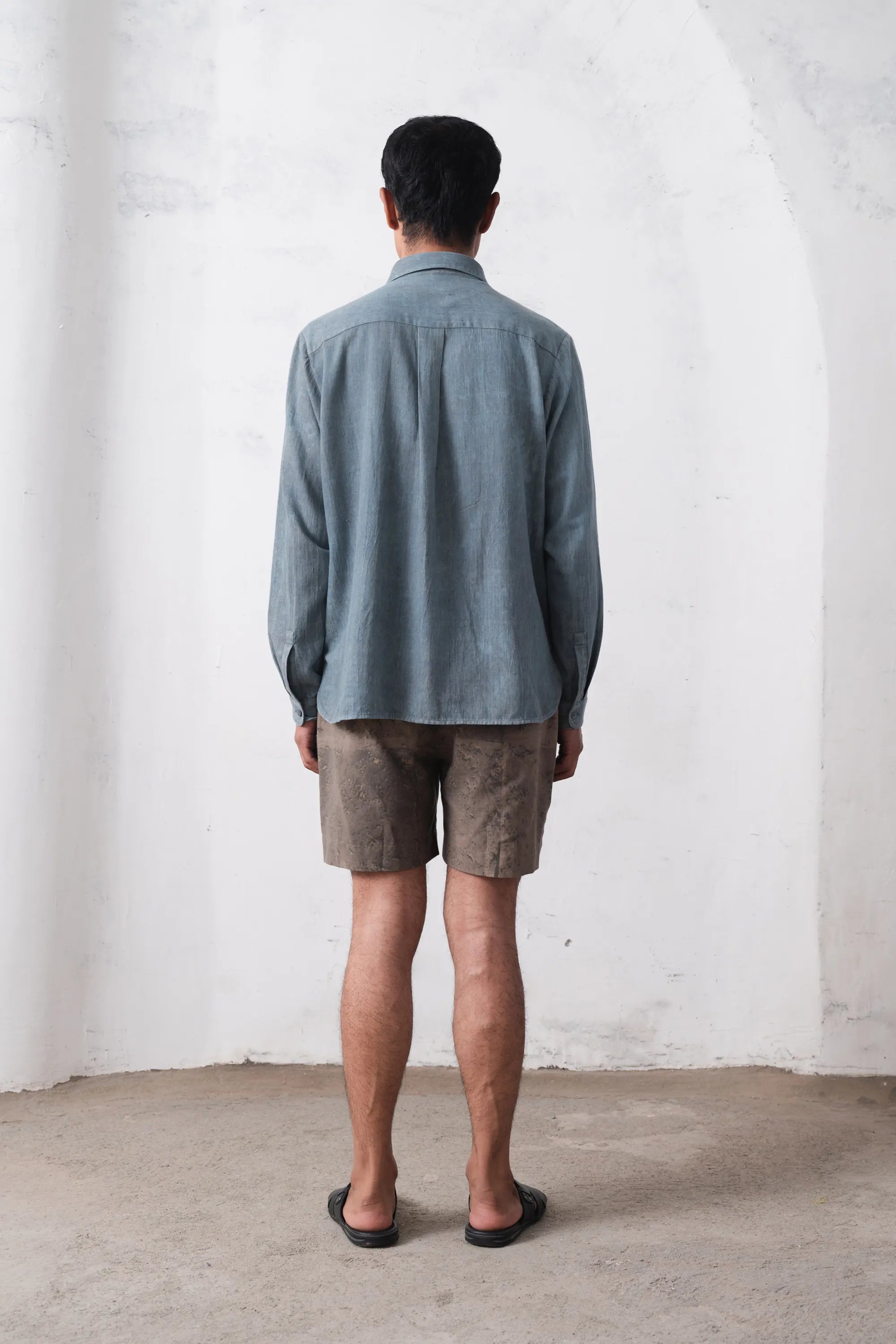Back shot of a person wearing a blue shirt and brown shorts standing against a white wall