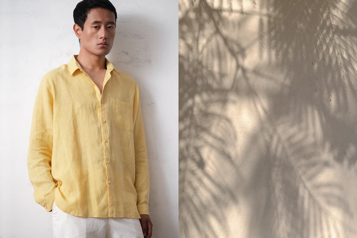 A Man wearing a yellow shirt standing against a white wall with a leafy shadow on the right.