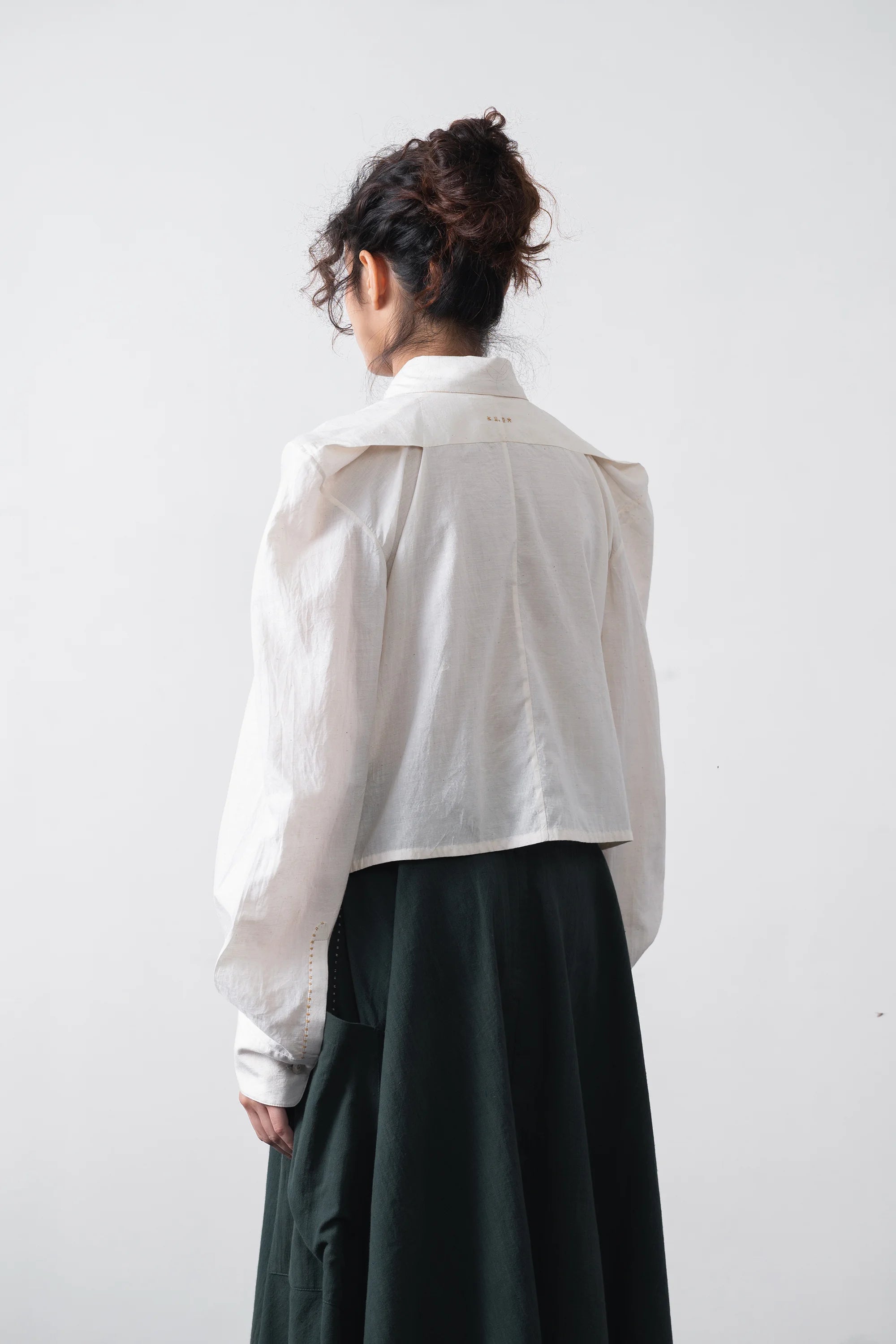 A woman shown from the back wearing a white cropped long-sleeve blouse with a wide collar and a skirt, photographed against a plain light grey studio background.