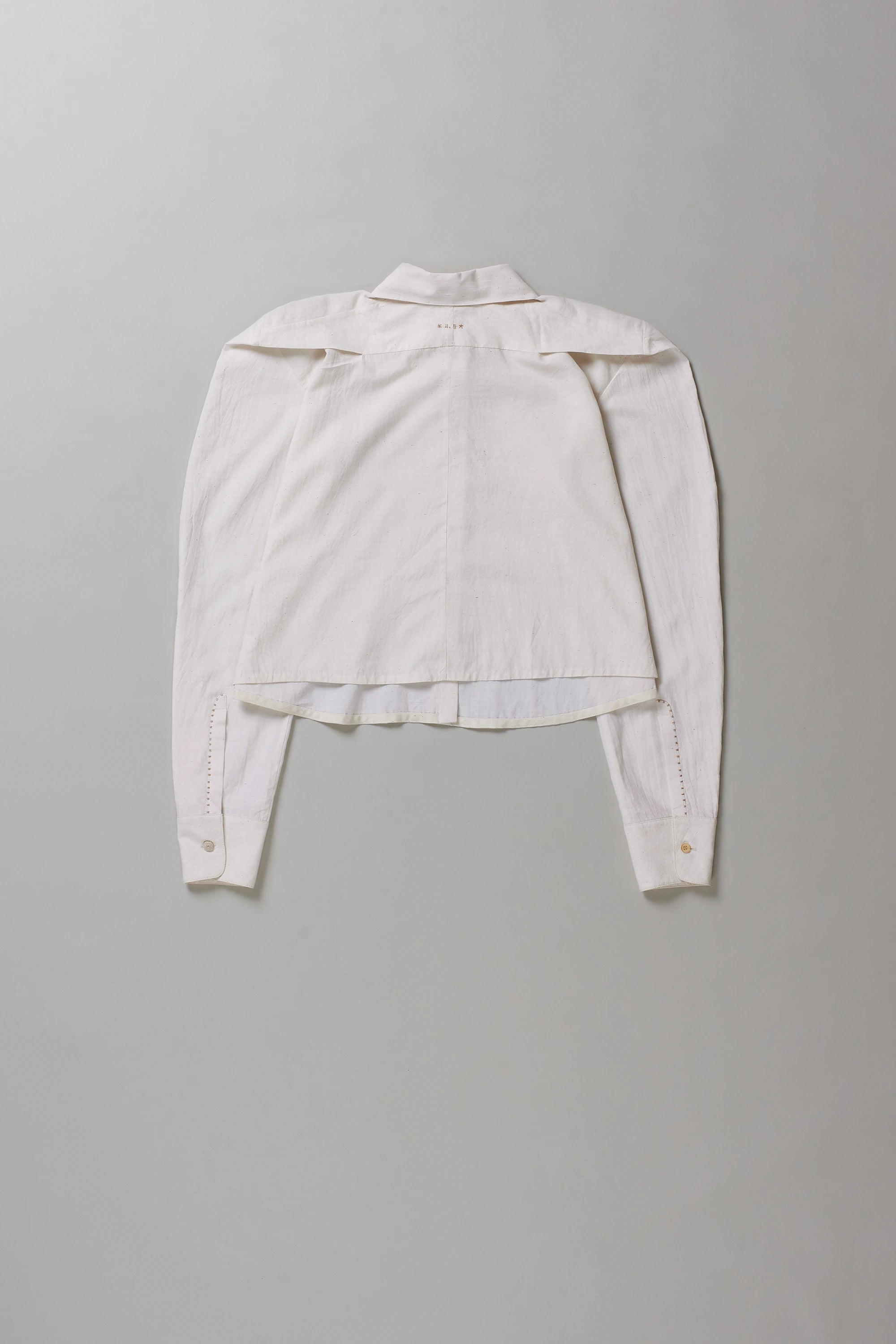 Back shot of off-white long-sleeve cropped shirt with structured shoulders and a high-low hem, laid flat against a neutral gray background.