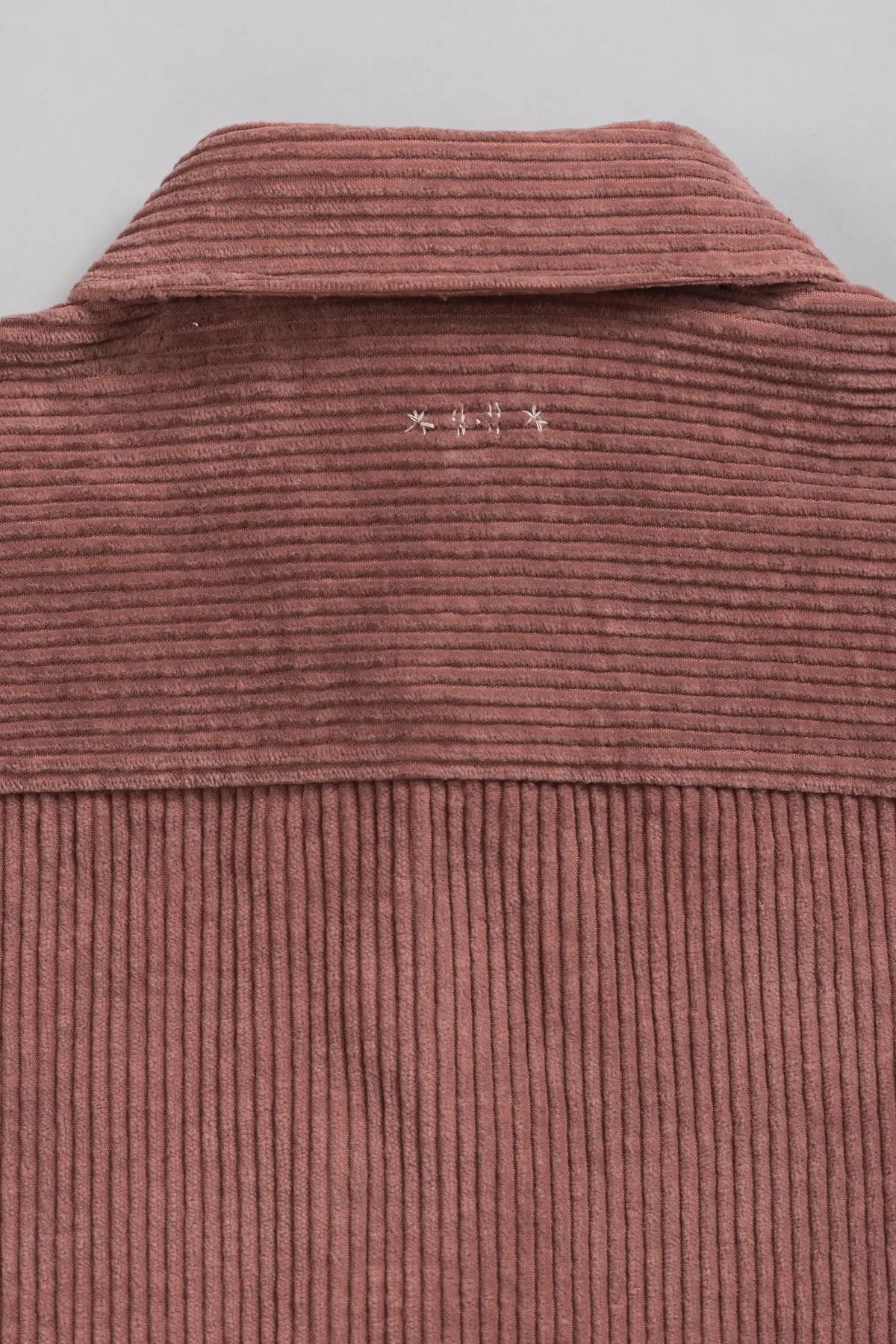 Close-up of a corduroy-like knit garment in a muted pink tone, showing the collar and vertical rib texture against a light grey background.