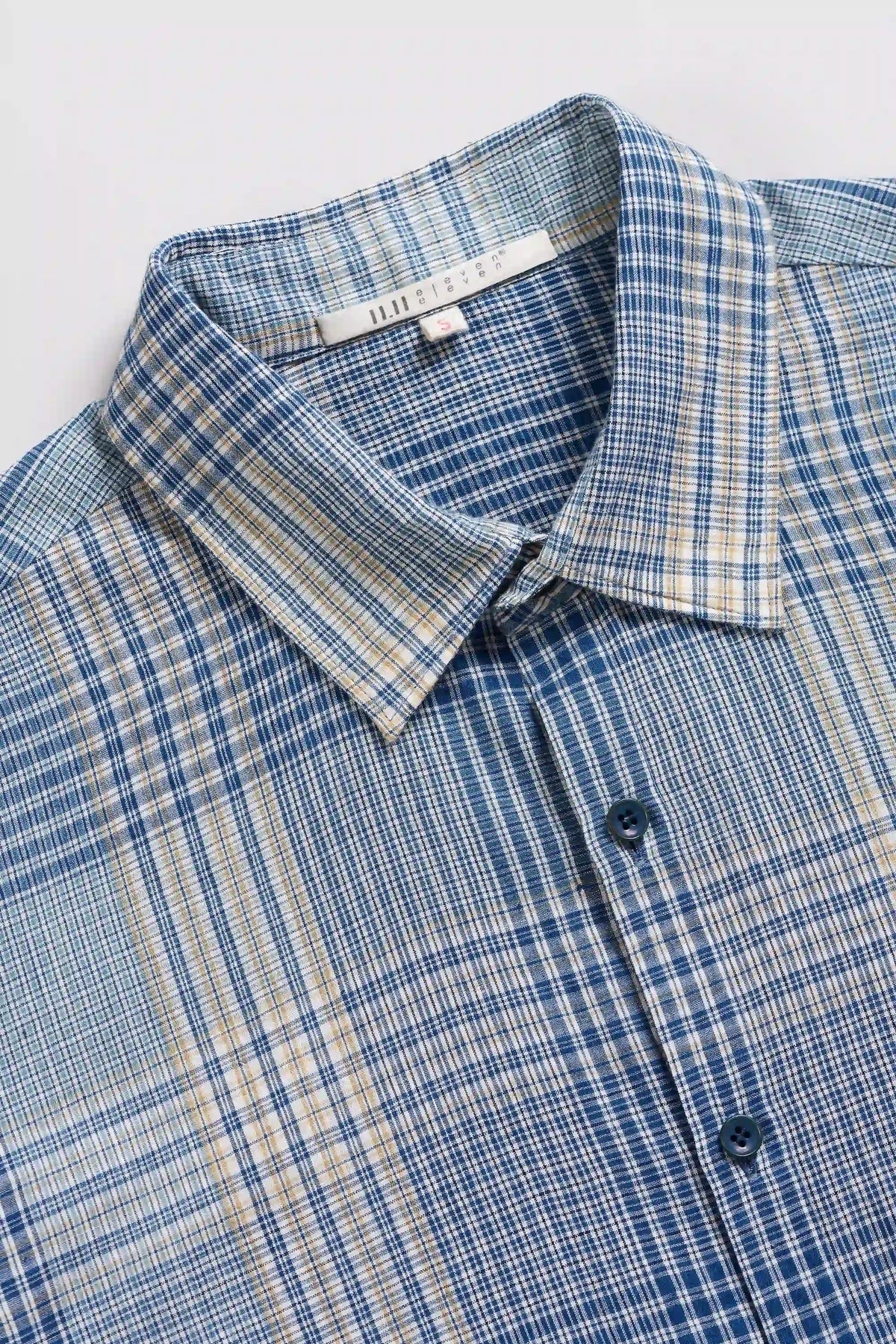 A close-up of a long-sleeve plaid button-down shirt in blue and beige, showing the collar, buttons, and fabric texture.