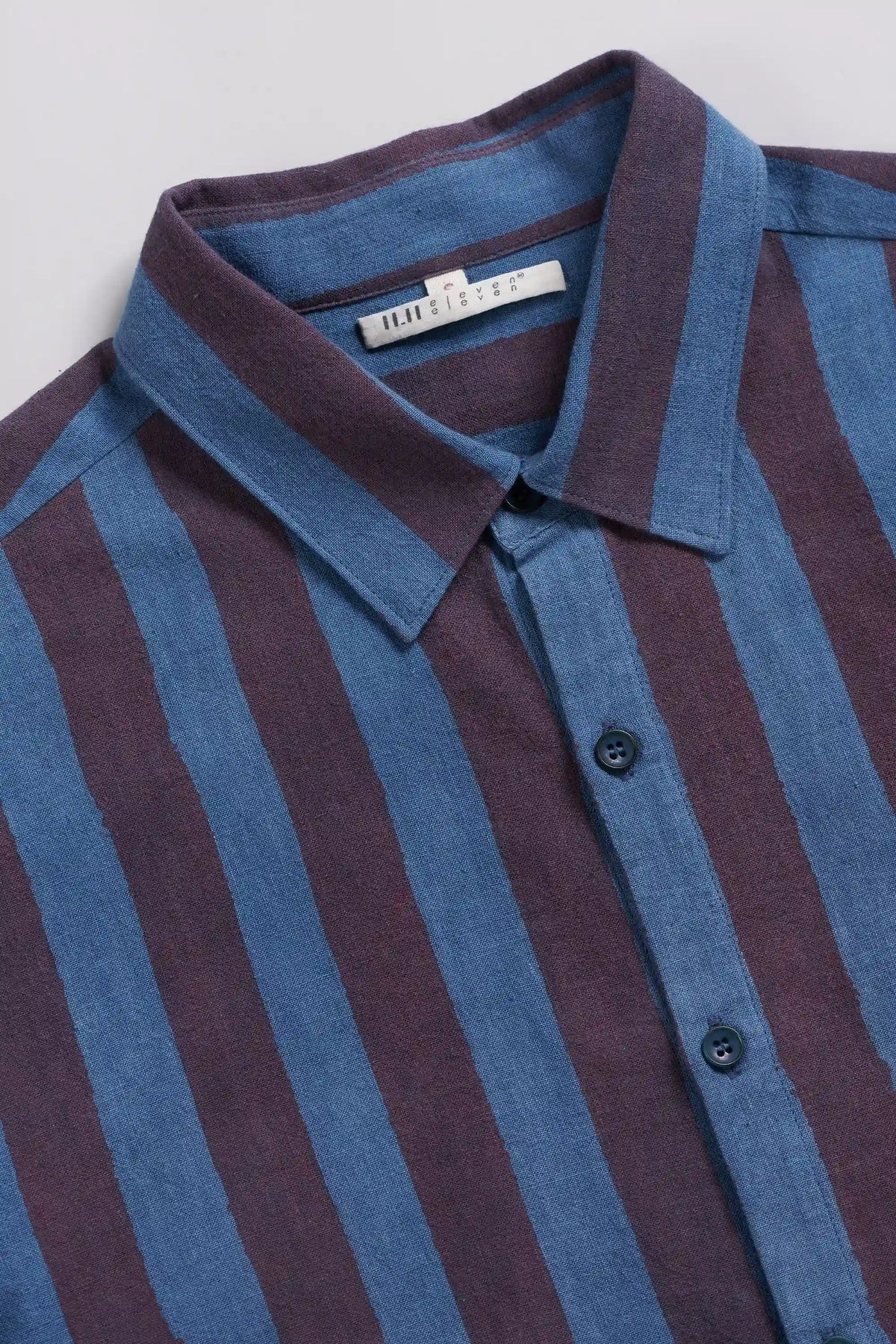 A close-up of a collared button-up shirt with vertical stripes in indigo and deep burgundy, featuring a white neck label that reads.