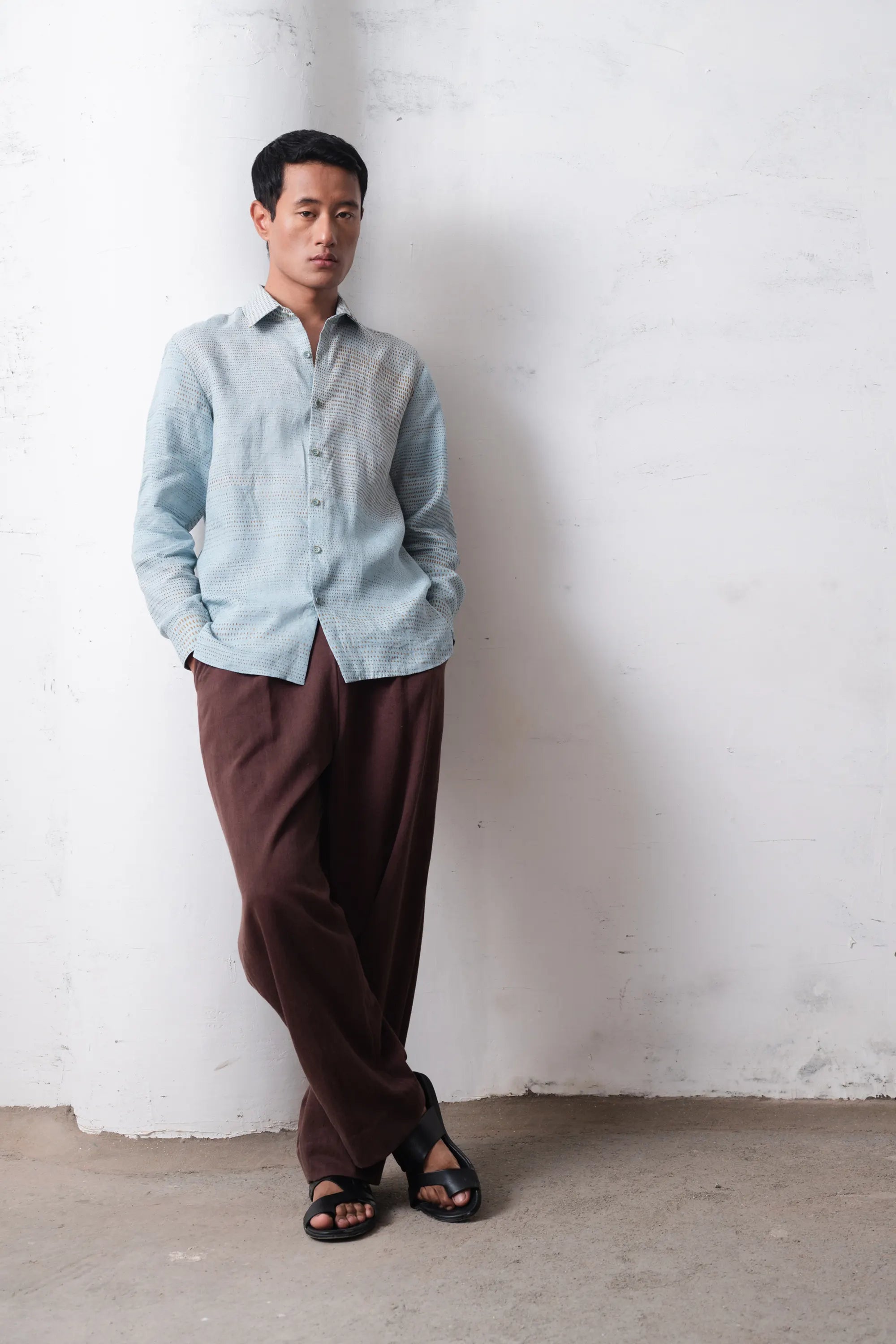 Man wearing a light blue shirt and brown pants against a white wall.
