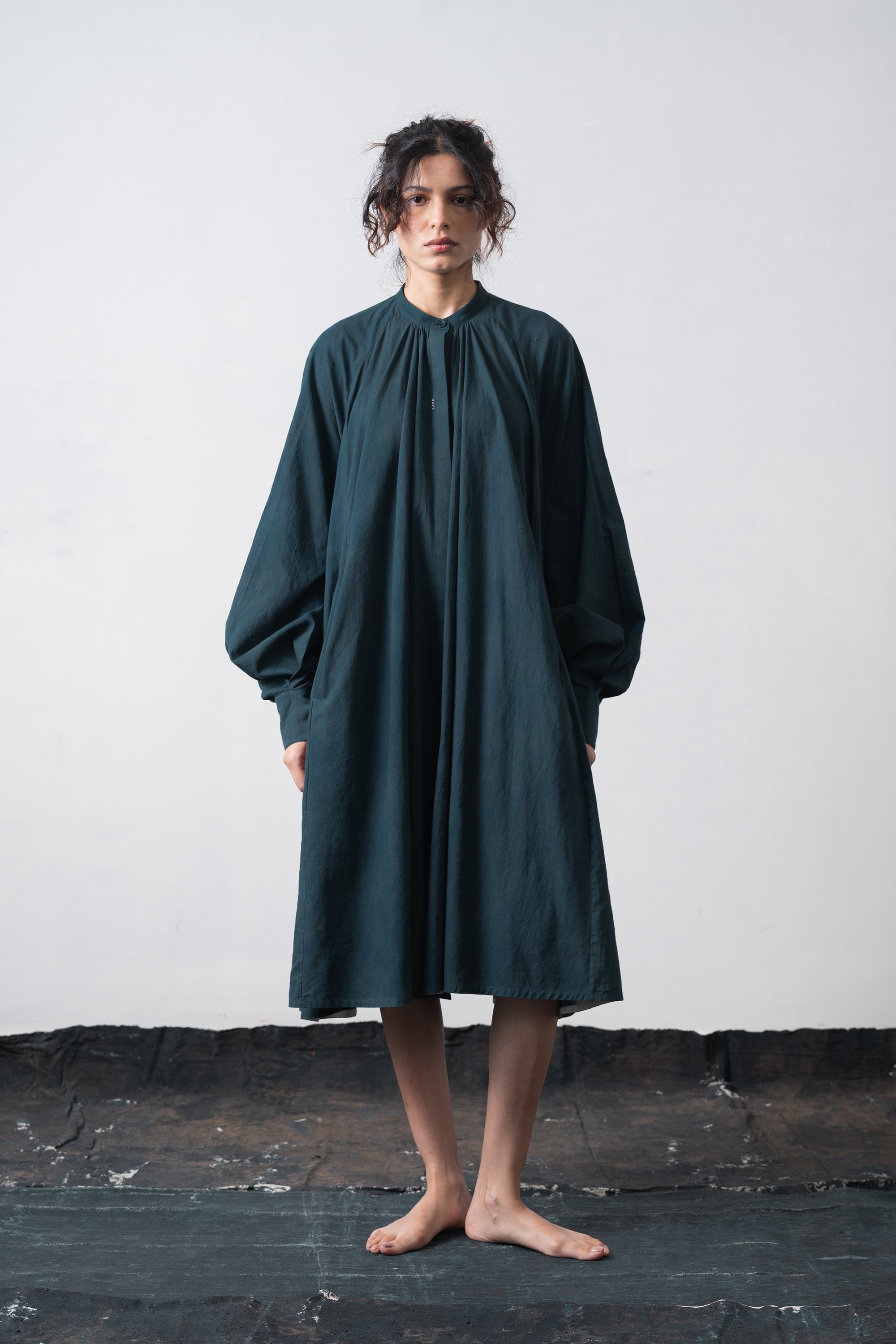 A woman stands barefoot against a wall wearing a loose, oversized black indigo dress with voluminous long sleeves and a gathered neckline.