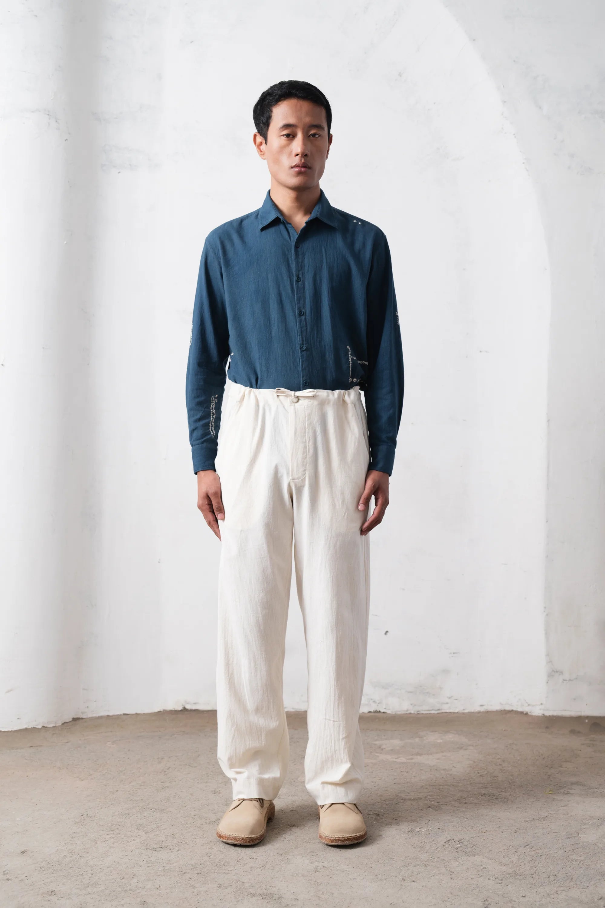 Man wearing a blue shirt and white pants standing against a light grey wall