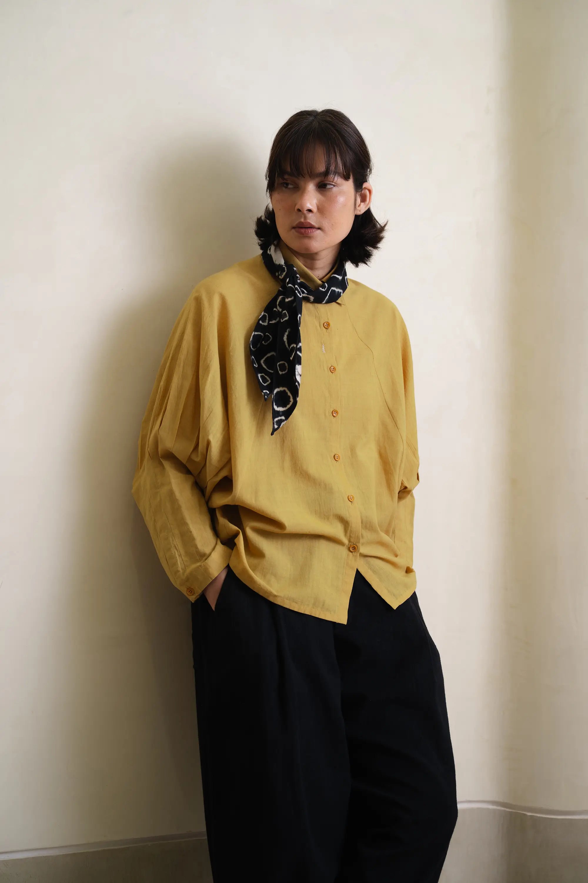 Person wearing a yellow shirt with a black patterned scarf, standing against a plain wall