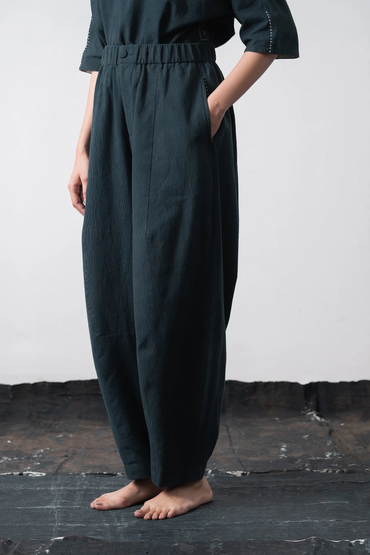A person wearing loose, black indigo wide-leg trousers with an elastic waistband and side pockets, standing barefoot against a grey wall and dark floor.