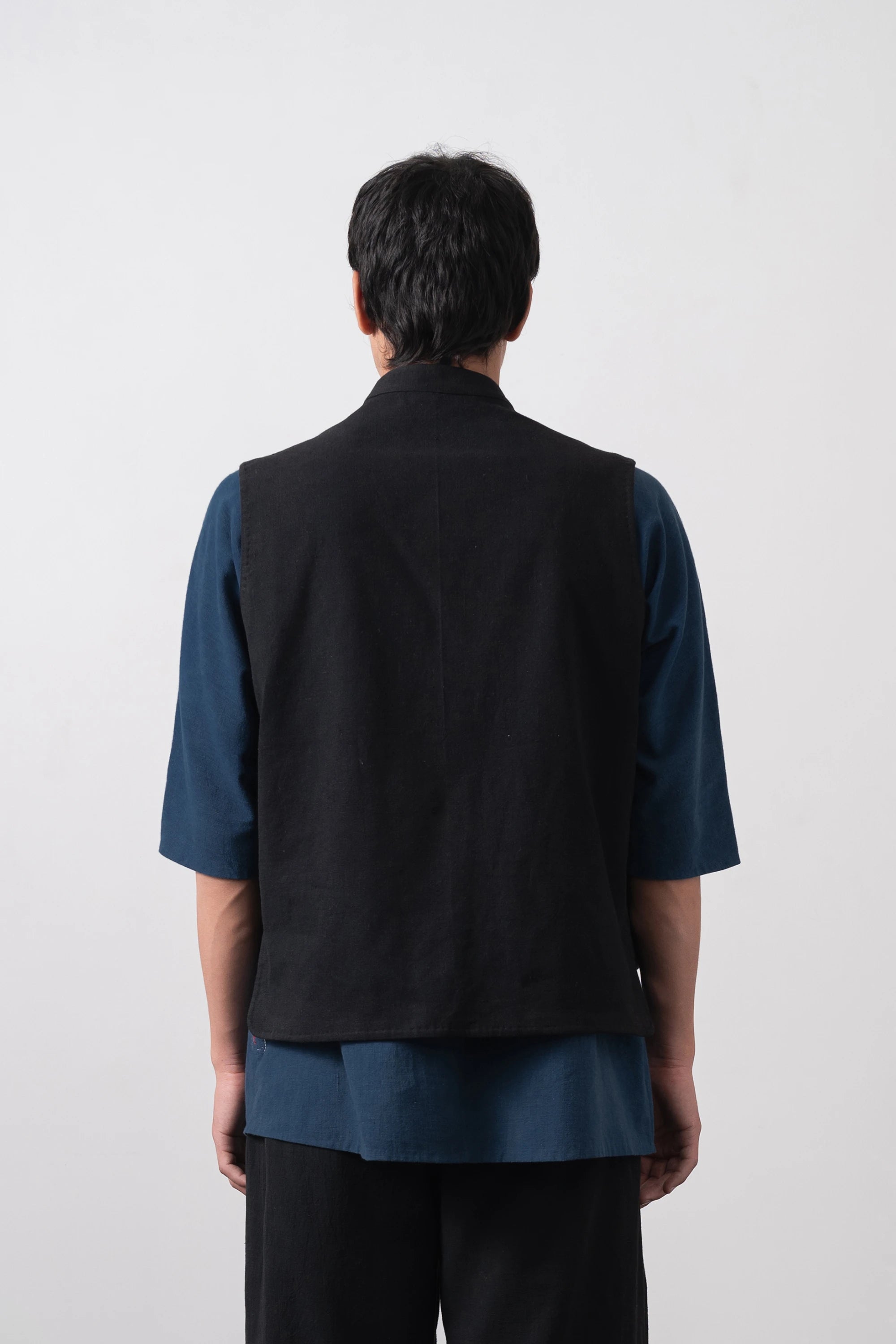 A man viewed from behind wearing a black sleeveless vest over a dark blue short-sleeve shirt and black pants, standing against a plain light grey background.