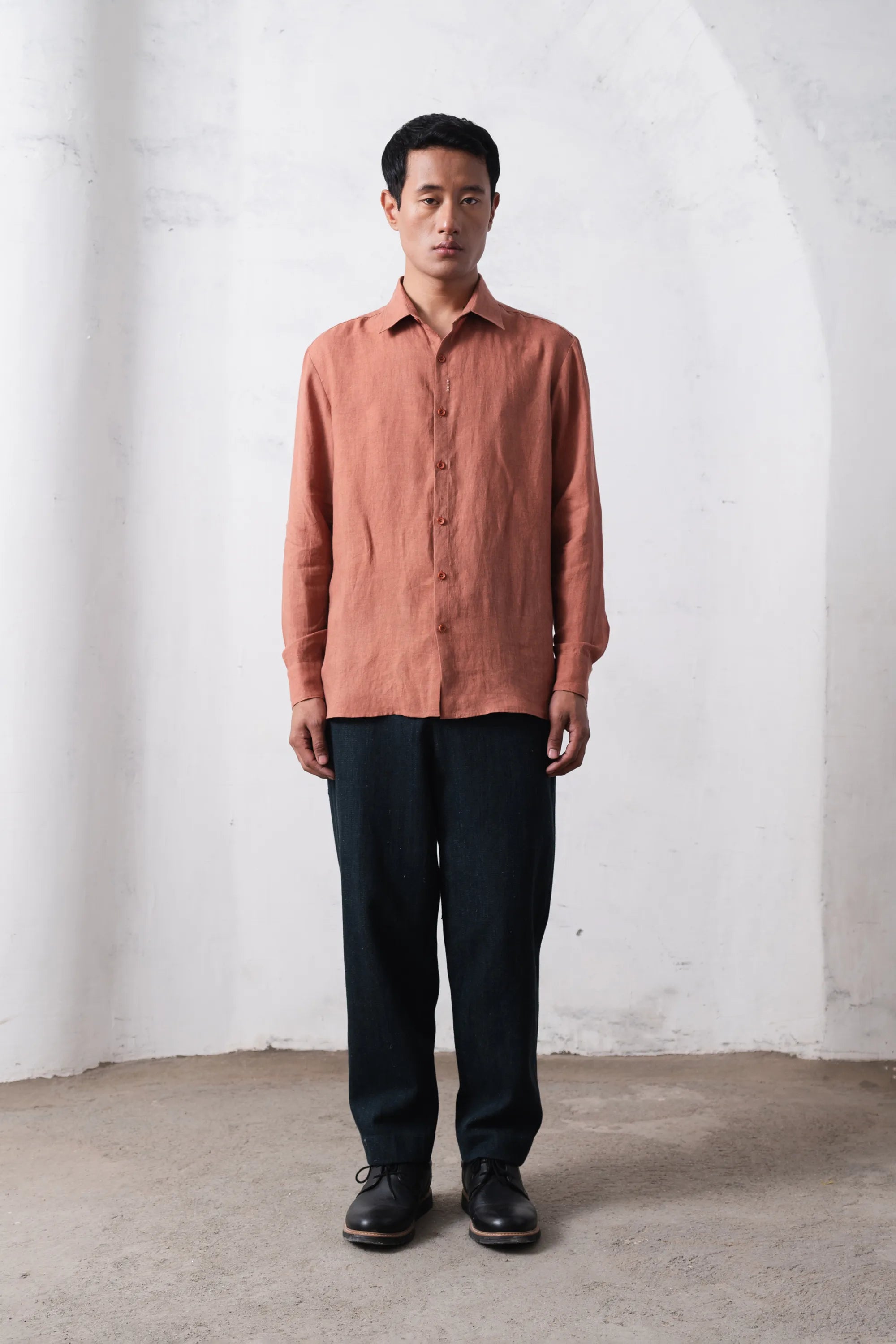 Man wearing a orange rust shirt and black pants against a light grey wall