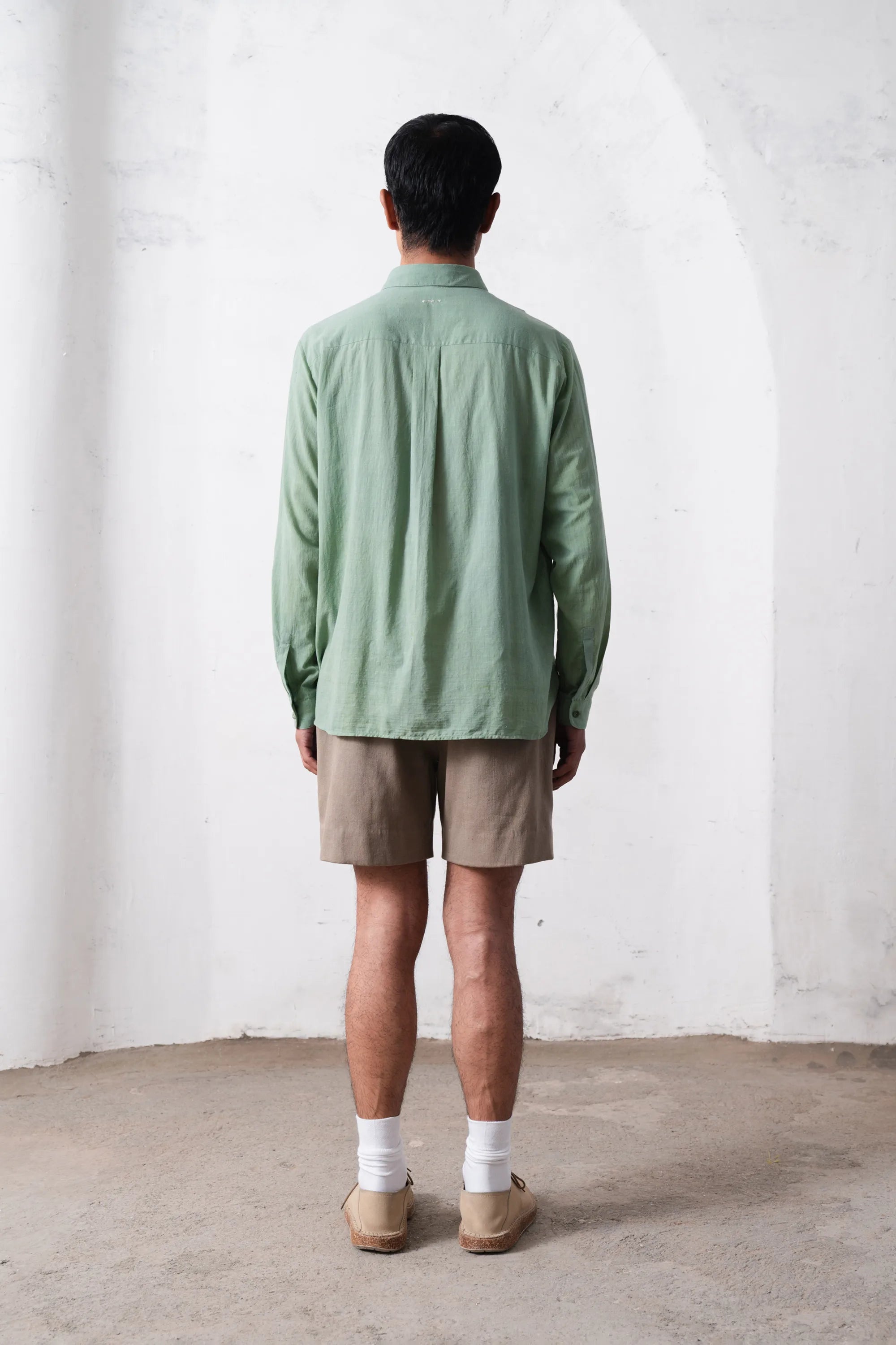 Back shot of a person wearing a turf green shirt and beige shorts standing against a light grey wall