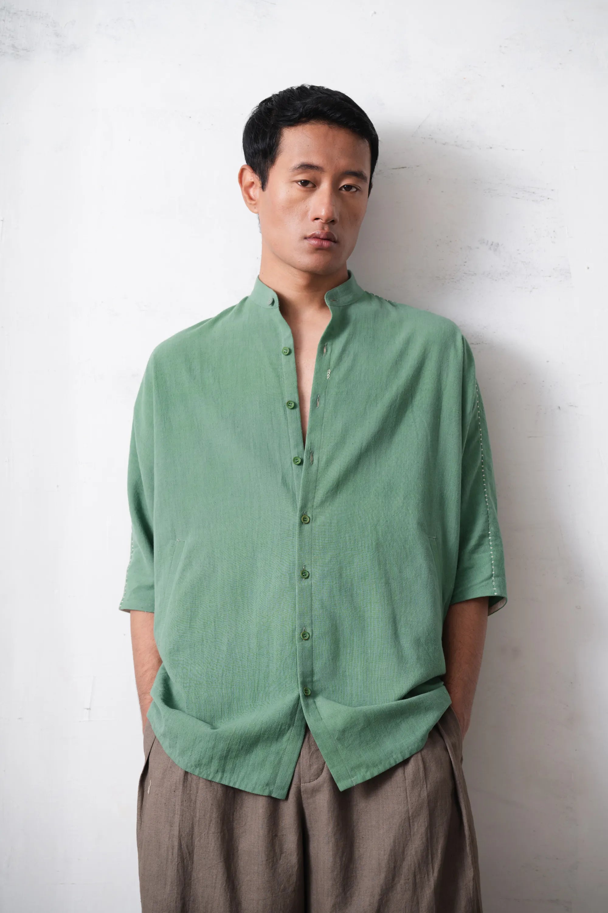 Man wearing a green shirt and brown pants against a light grey wall
