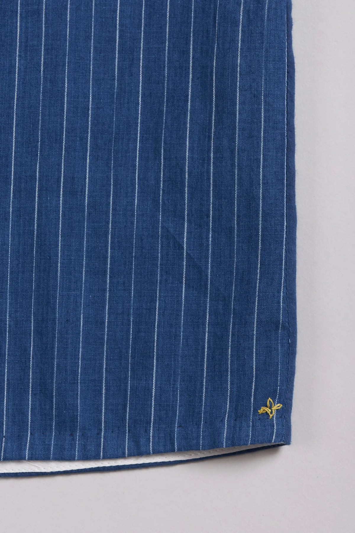 A close-up of a indigo fabric, featuring thin white vertical pinstripes and a small yellow embroidered near the bottom right corner, set against a light grey background.