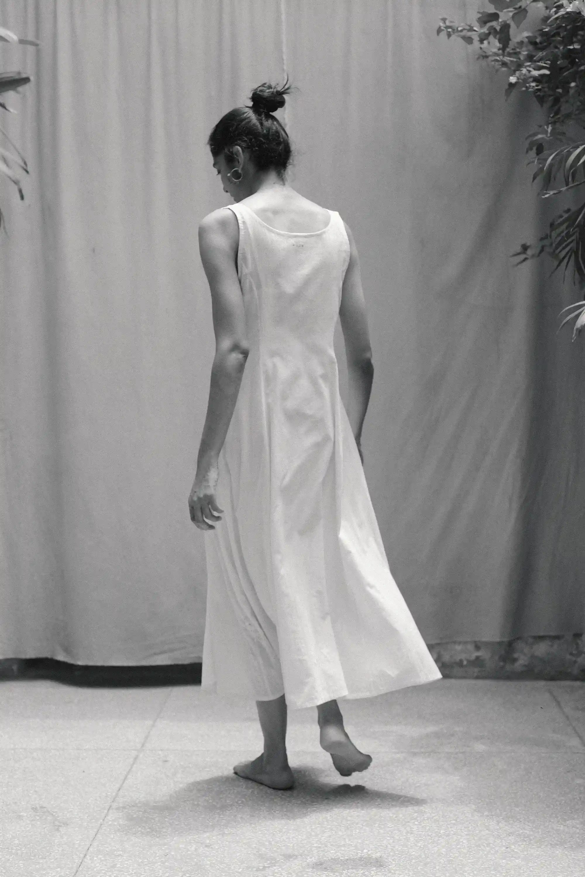 Back shot of a woman wearing a ecru dress standing against a plain background with plants.