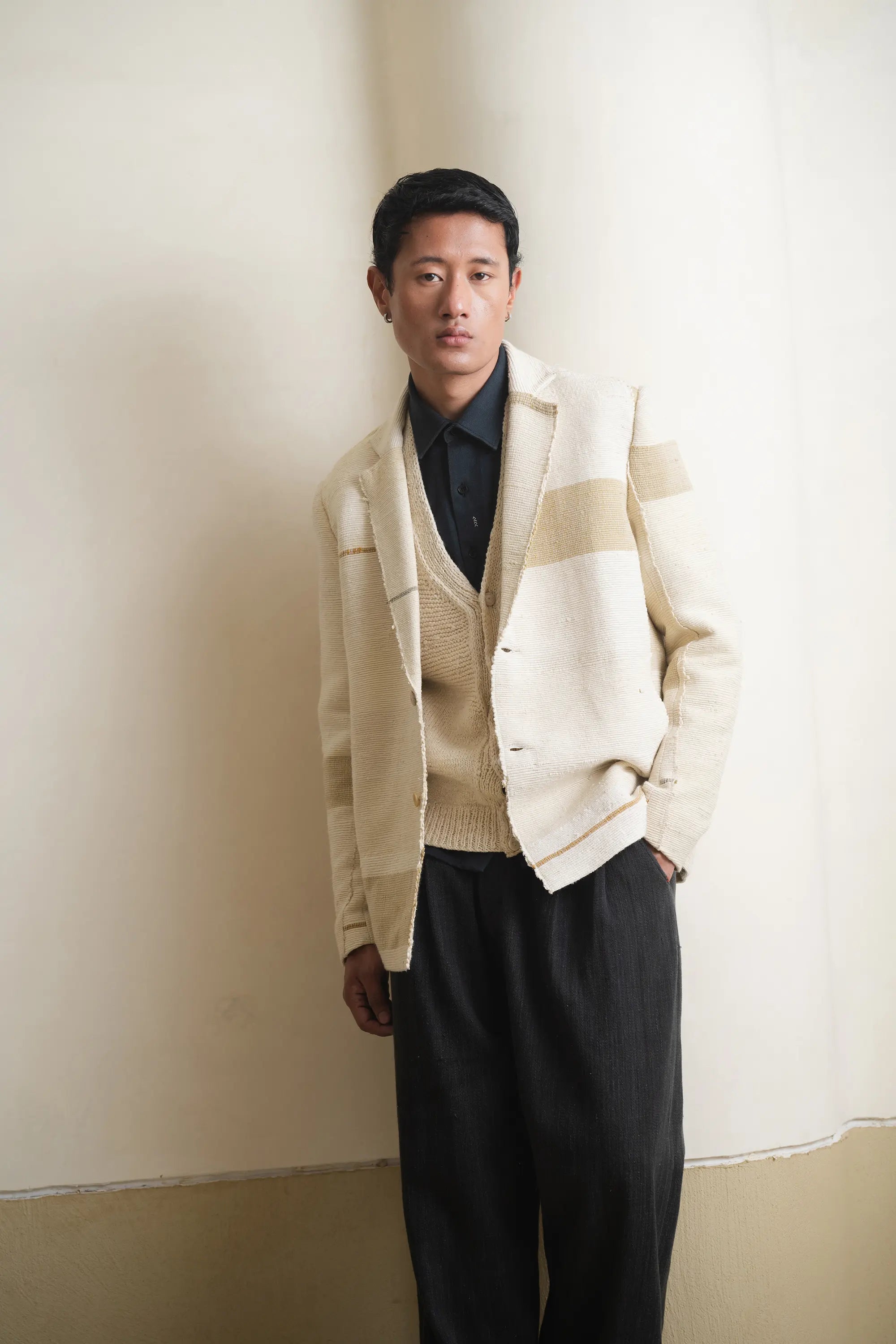 Man wearing a beige cardigan over a black shirt and pants against a light grey wall