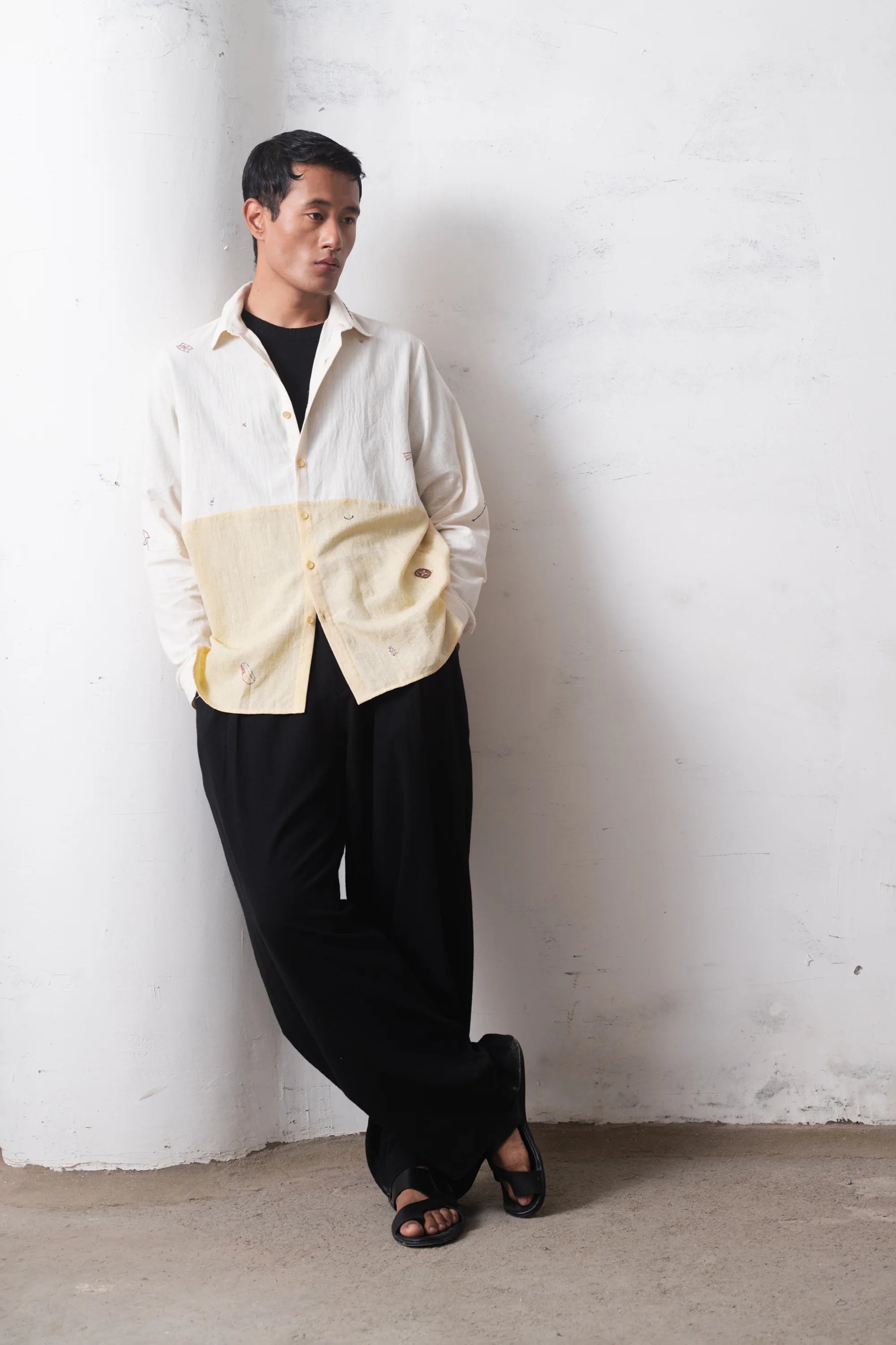 Man wearing a white and yellow shirt with black pants against a light grey wall