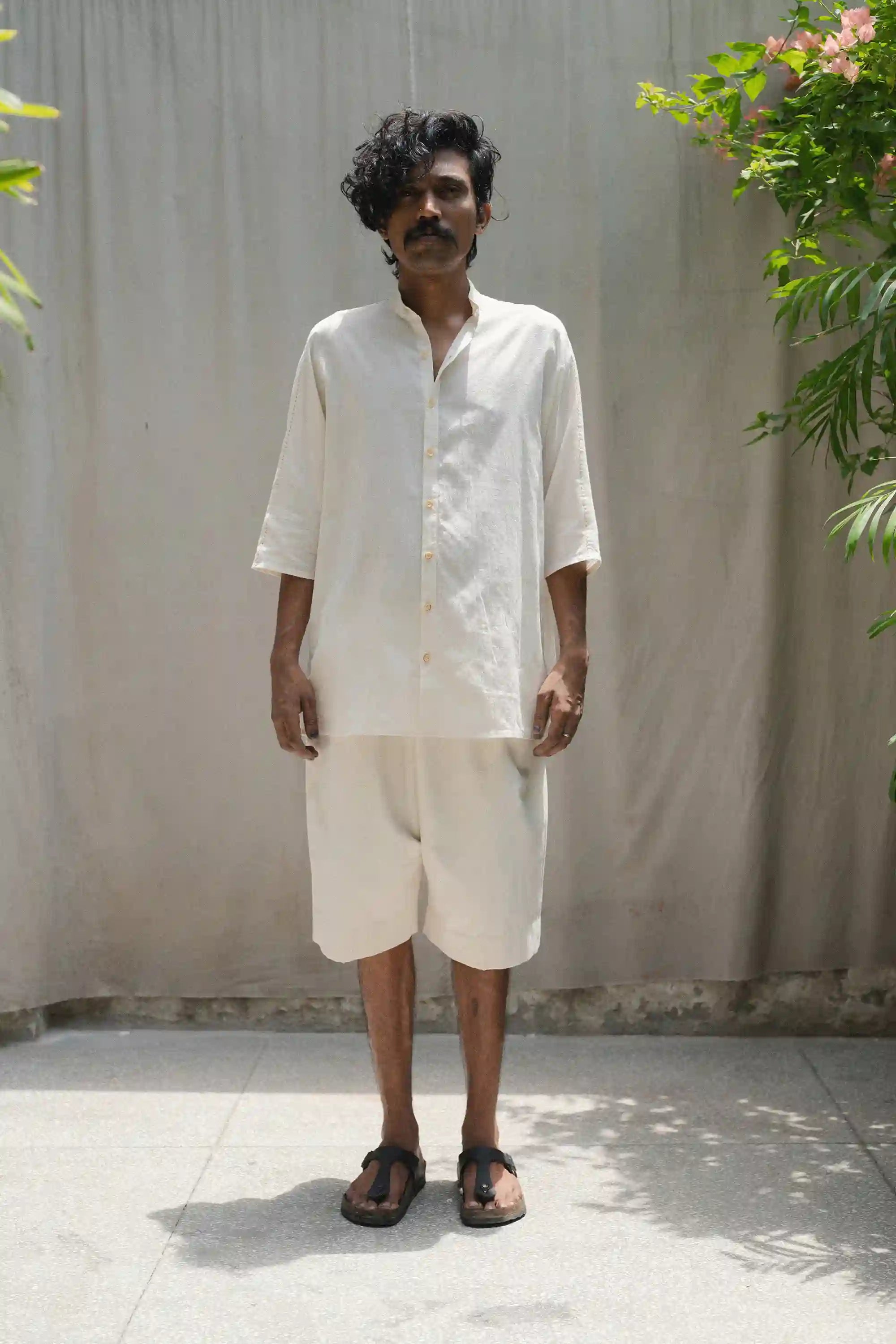 Man wearing a ecru shirt and shorts standing against a plain wall with plants around.
