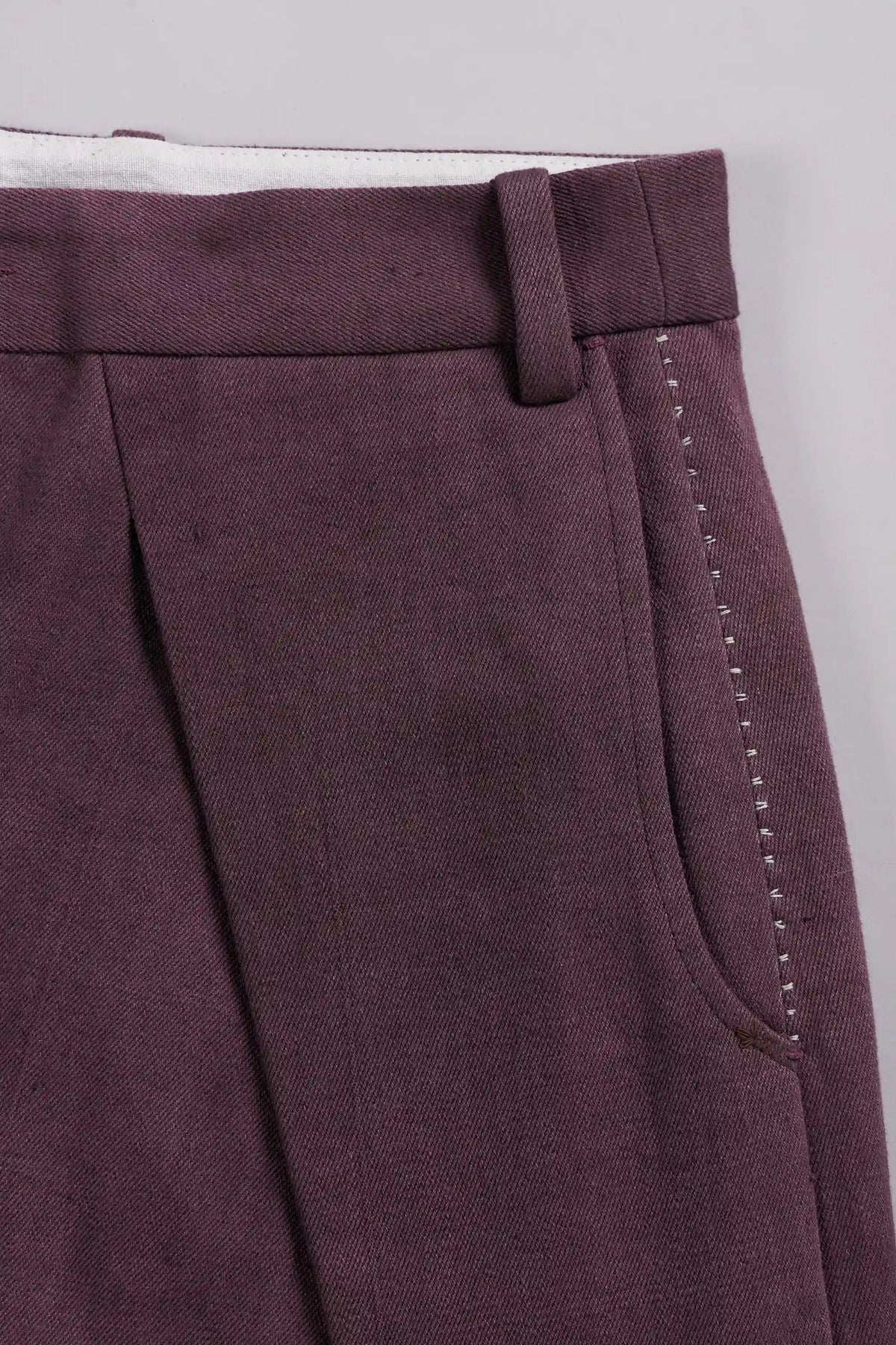Close-up of a pair of pleated trousers in a purple red fabric, showing the waistband, belt loop, front pleat, and side pocket with decorative stitching.