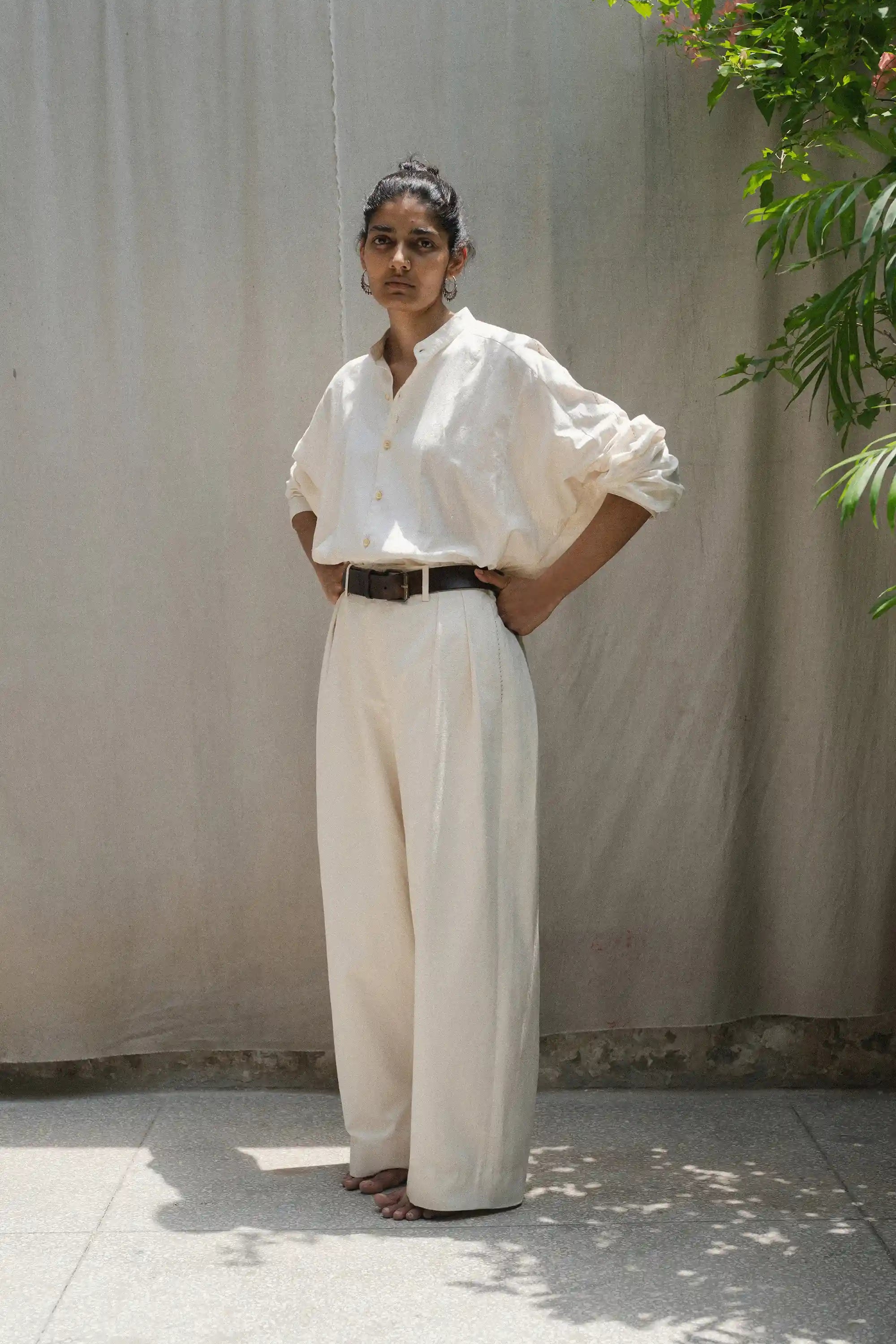 Person wearing a white shirt and ecru pants standing against a plain wall with greenery.