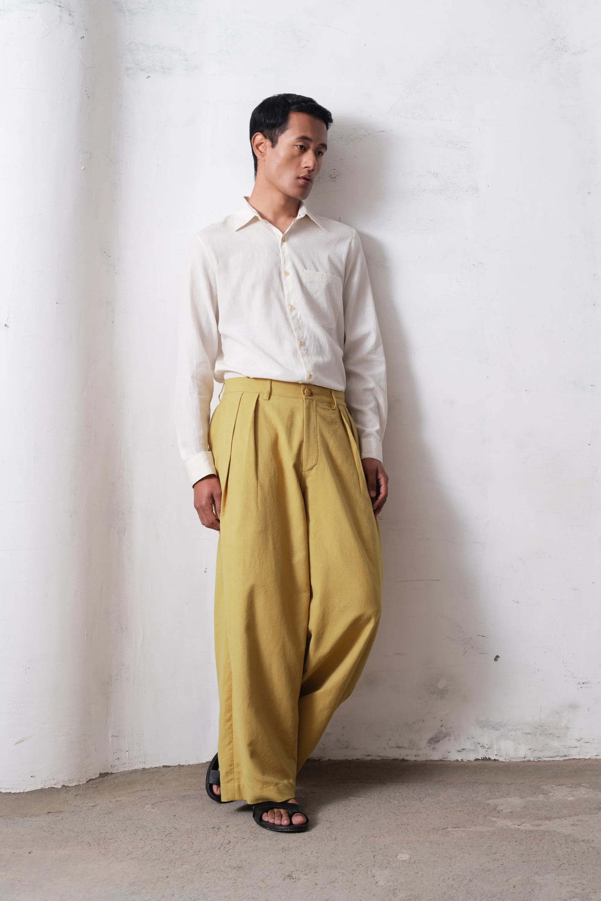 Person wearing a white shirt and ochre yellow pants against a white wall
