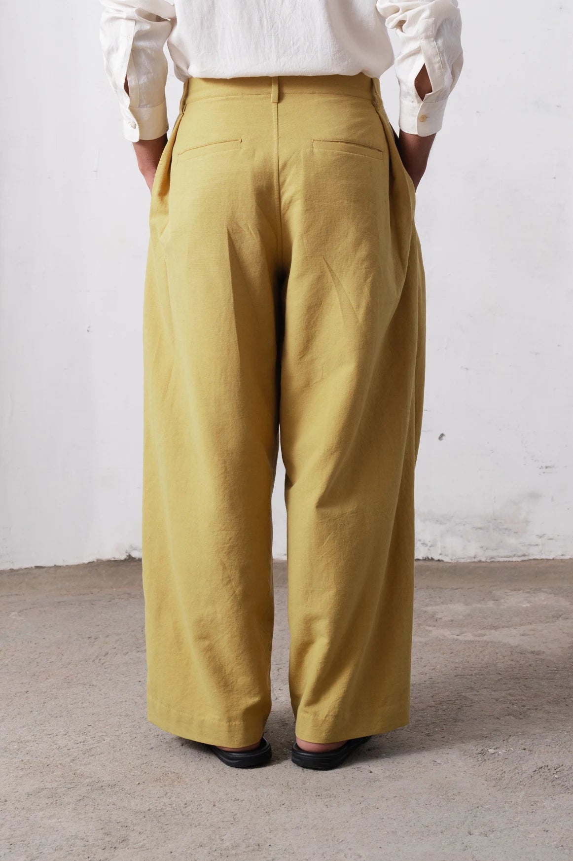 Back shot of a person wearing ochre yellow pants and a white shirt against a light grey background