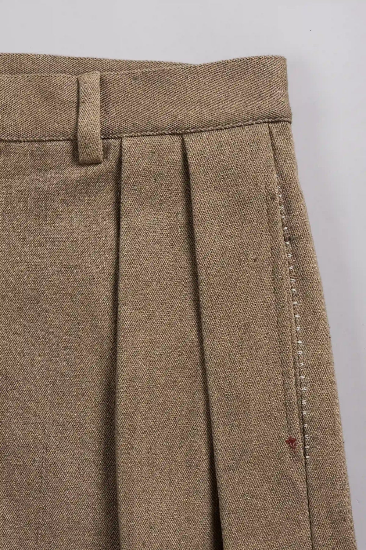 Close-up of pleated trousers in a textured taupe-brown fabric, showing the waistband, belt loop, and side stitching.